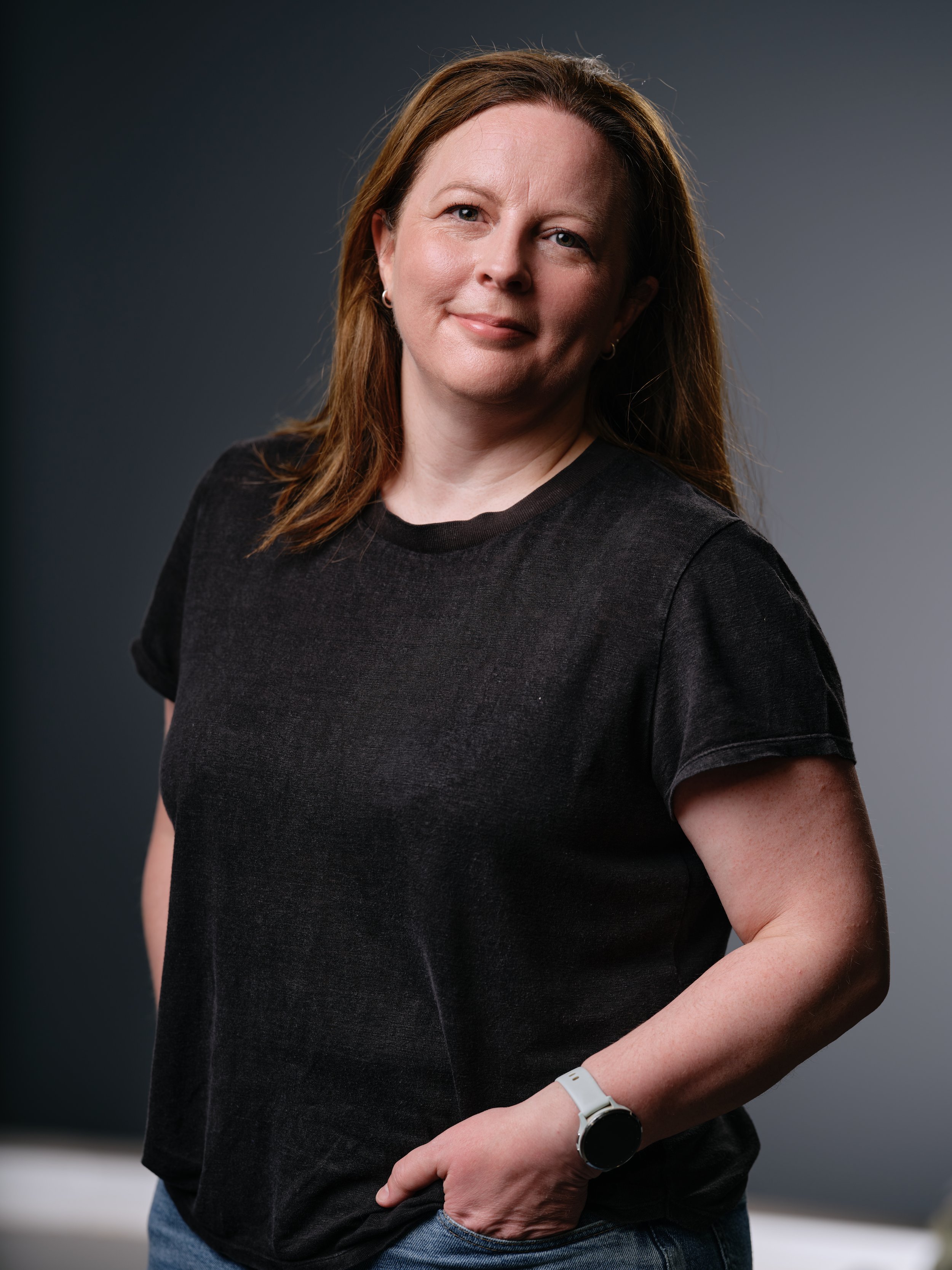 A woman with light skin and shoulder-length brown hair, wearing a black t-shirt and a watch on her left wrist, standing against a dark background.
