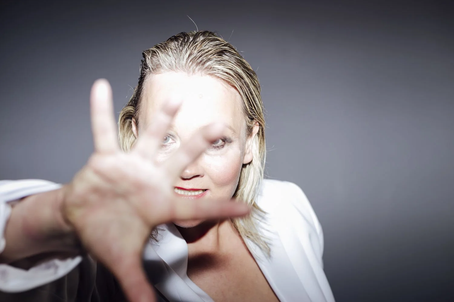 A woman with blonde hair reaching toward the camera with her hand, partially obscuring her face, against a dark gray background.