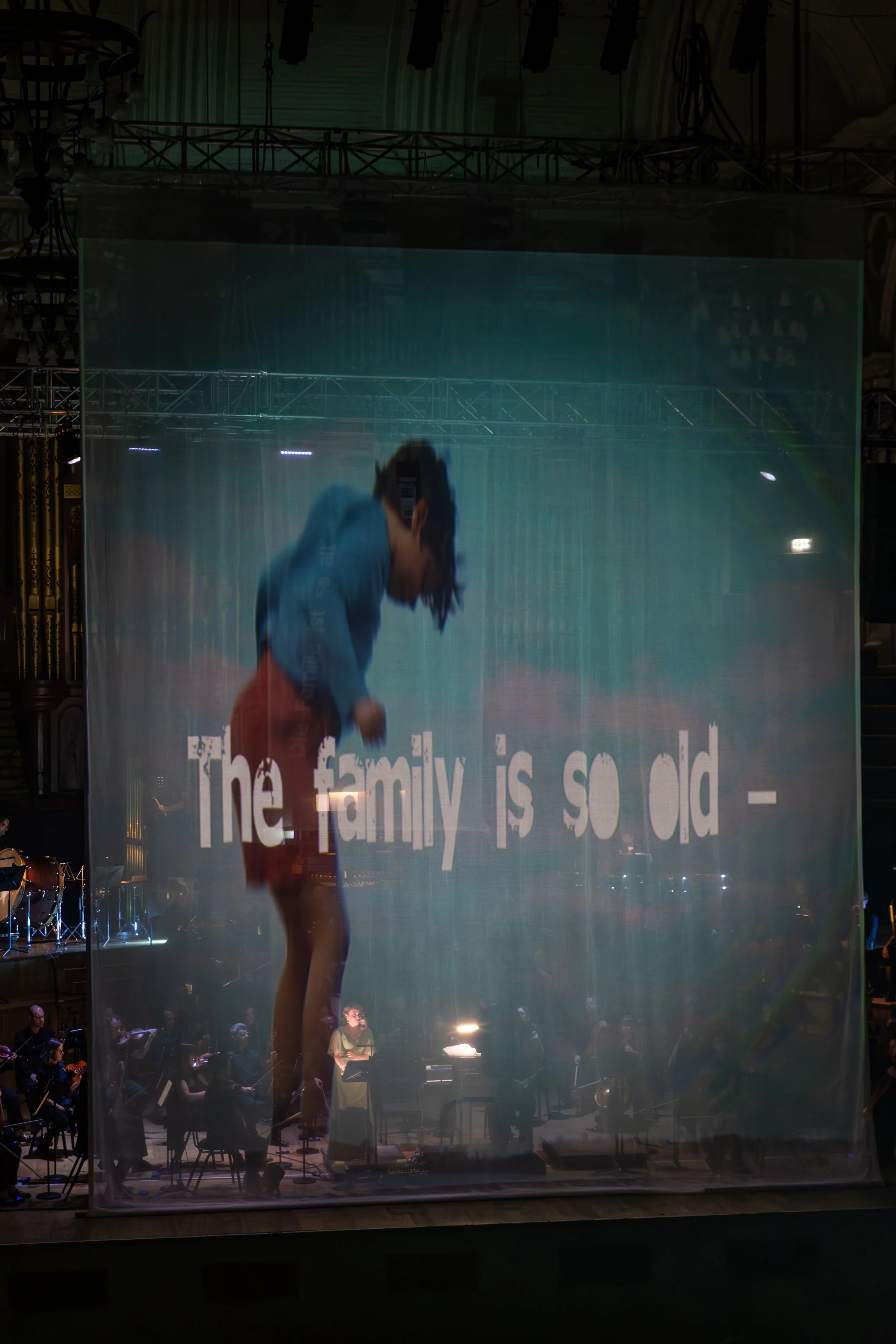 A person in a blue shirt and orange shorts bending forward, standing on stage in front of an orchestra, with a projection screen displaying the partially visible white text 'The family is so old -' behind them.