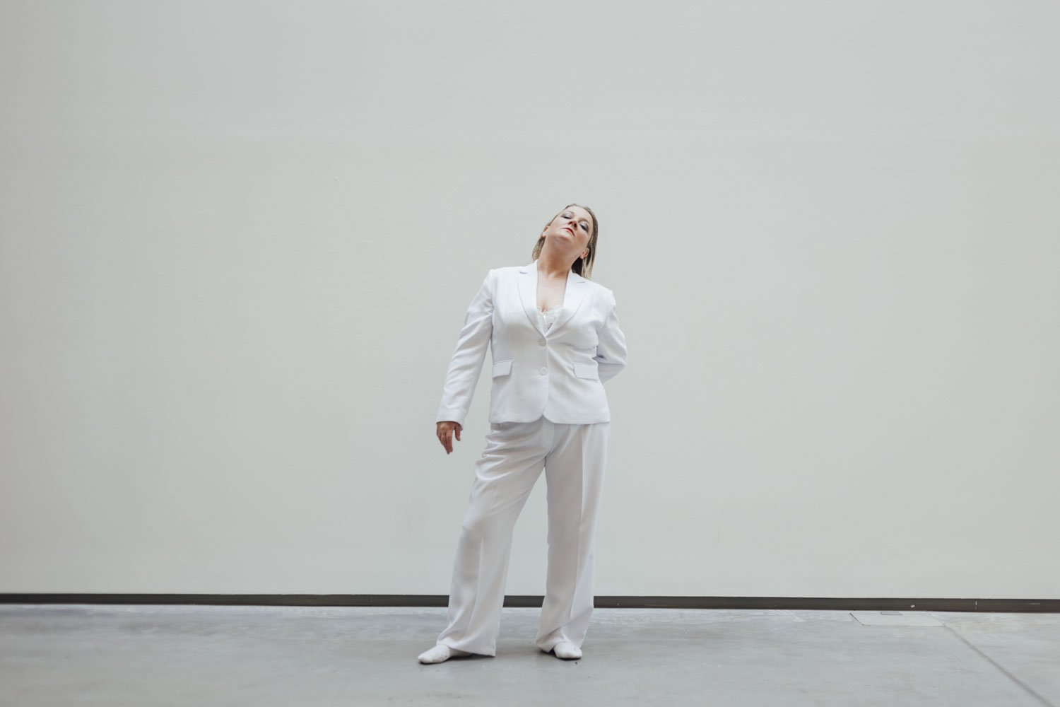 A woman in an all-white suit standing against a plain light gray wall, with her head tilted back and eyes closed.
