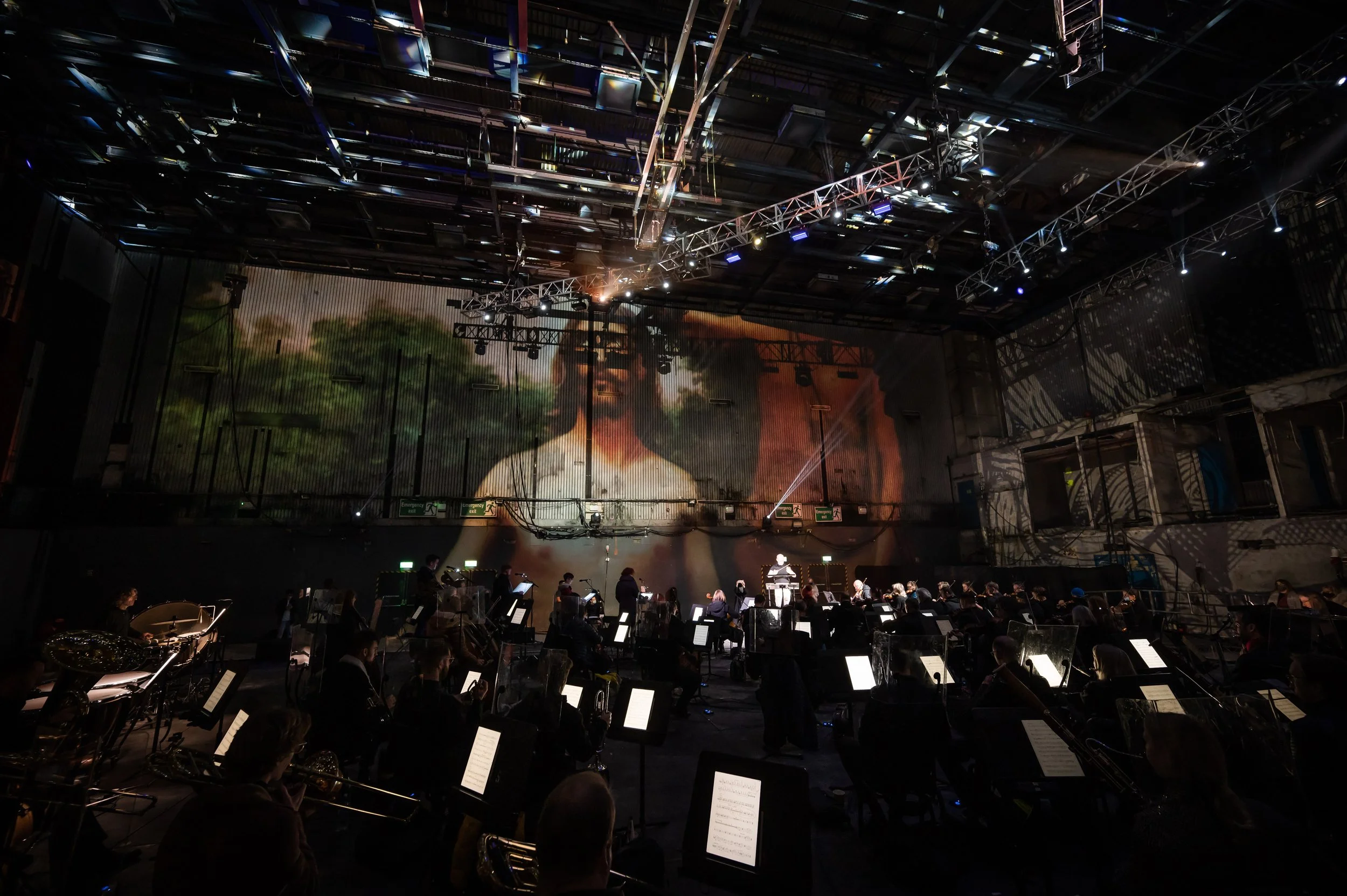 Orchestra performing on stage in a large indoor concert hall with stage lighting and a screen projecting an image in the background.