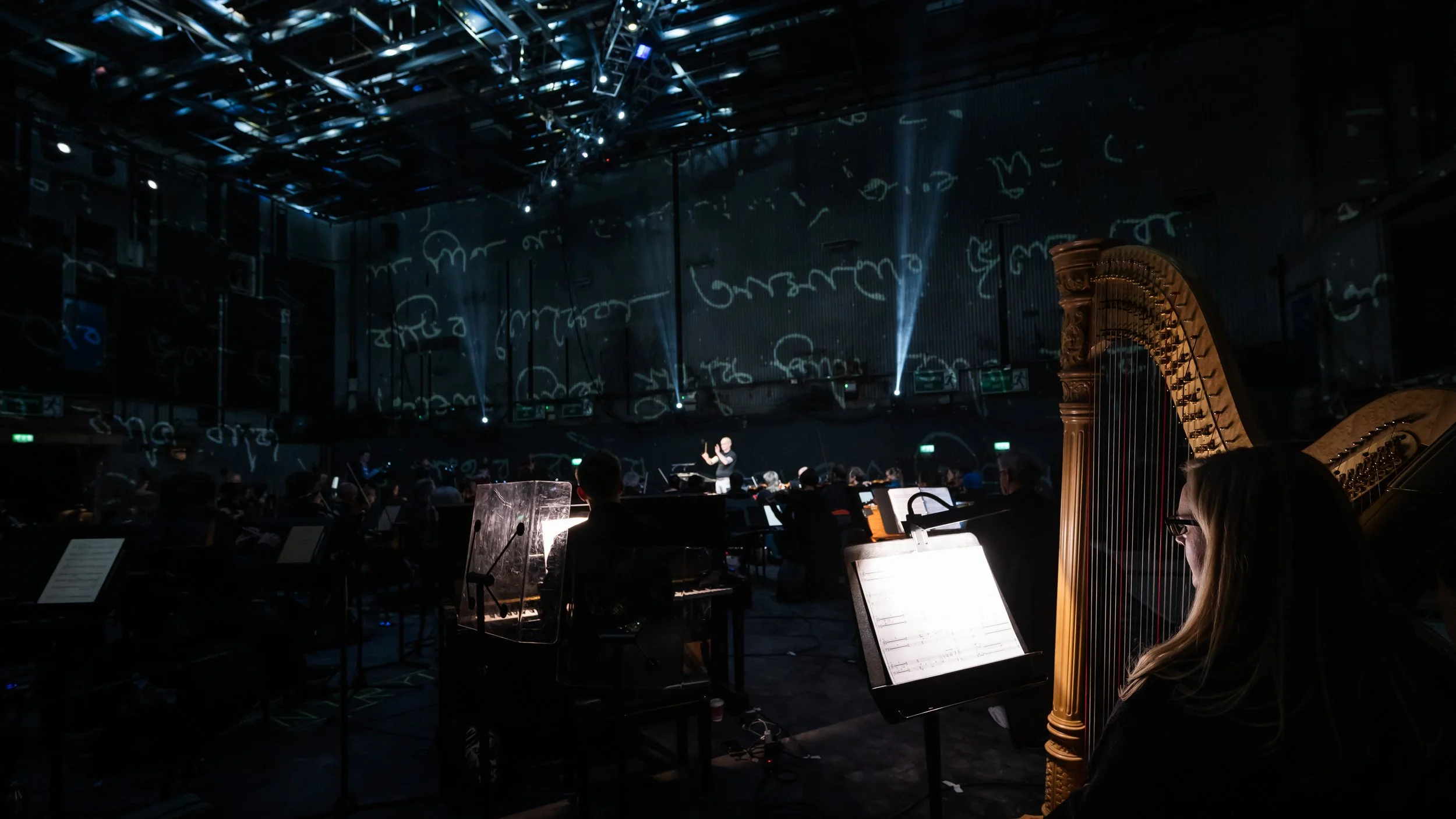 An orchestra performing on stage with a conductor, a grand harp on the right, and musical scores on music stands in a dimly lit concert hall with blue lighting and abstract projections on the walls.