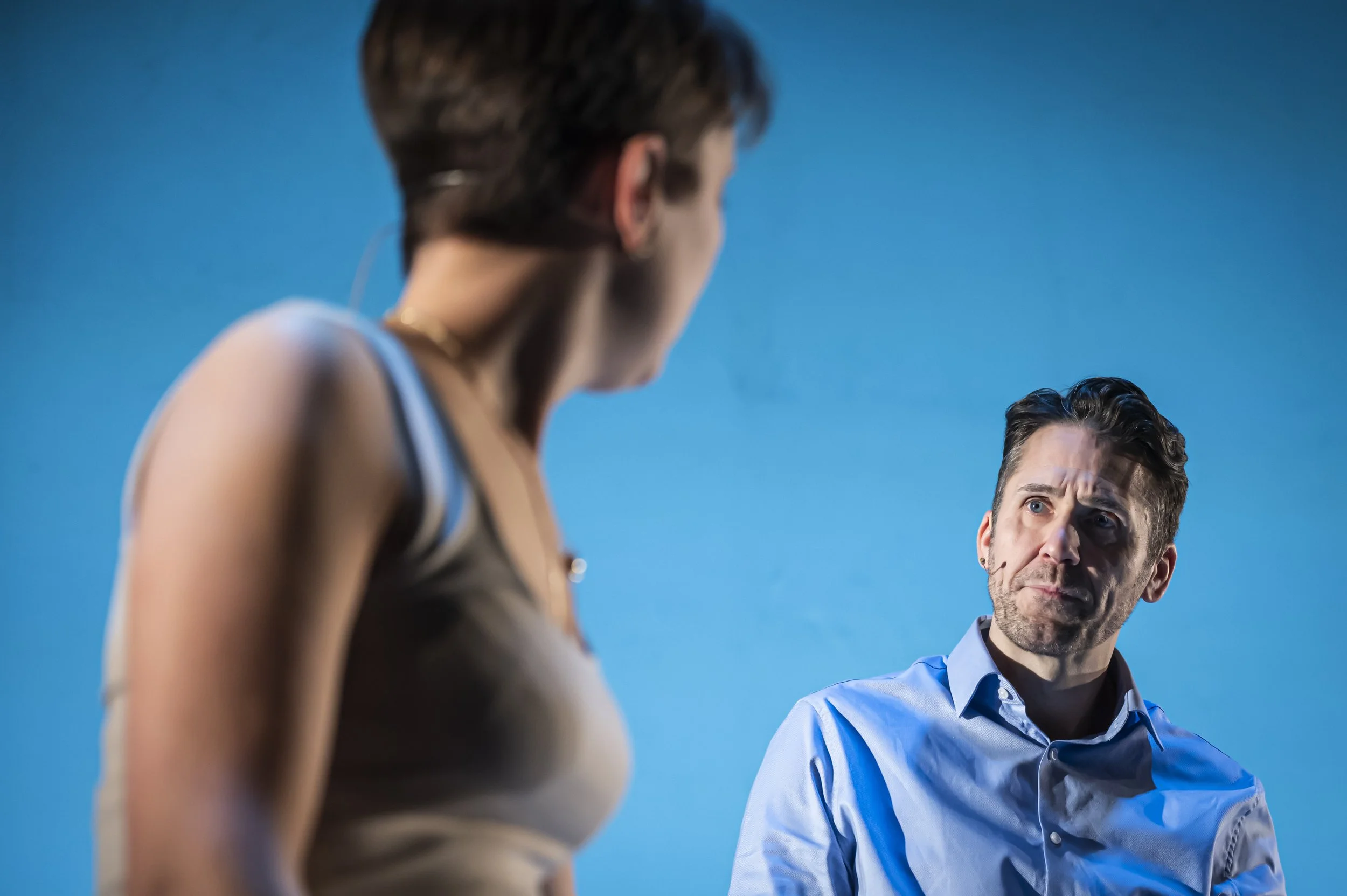 A woman and a man appear to be engaged in a serious conversation, with the woman facing away from the camera and the man looking intently at her against a clear blue sky.