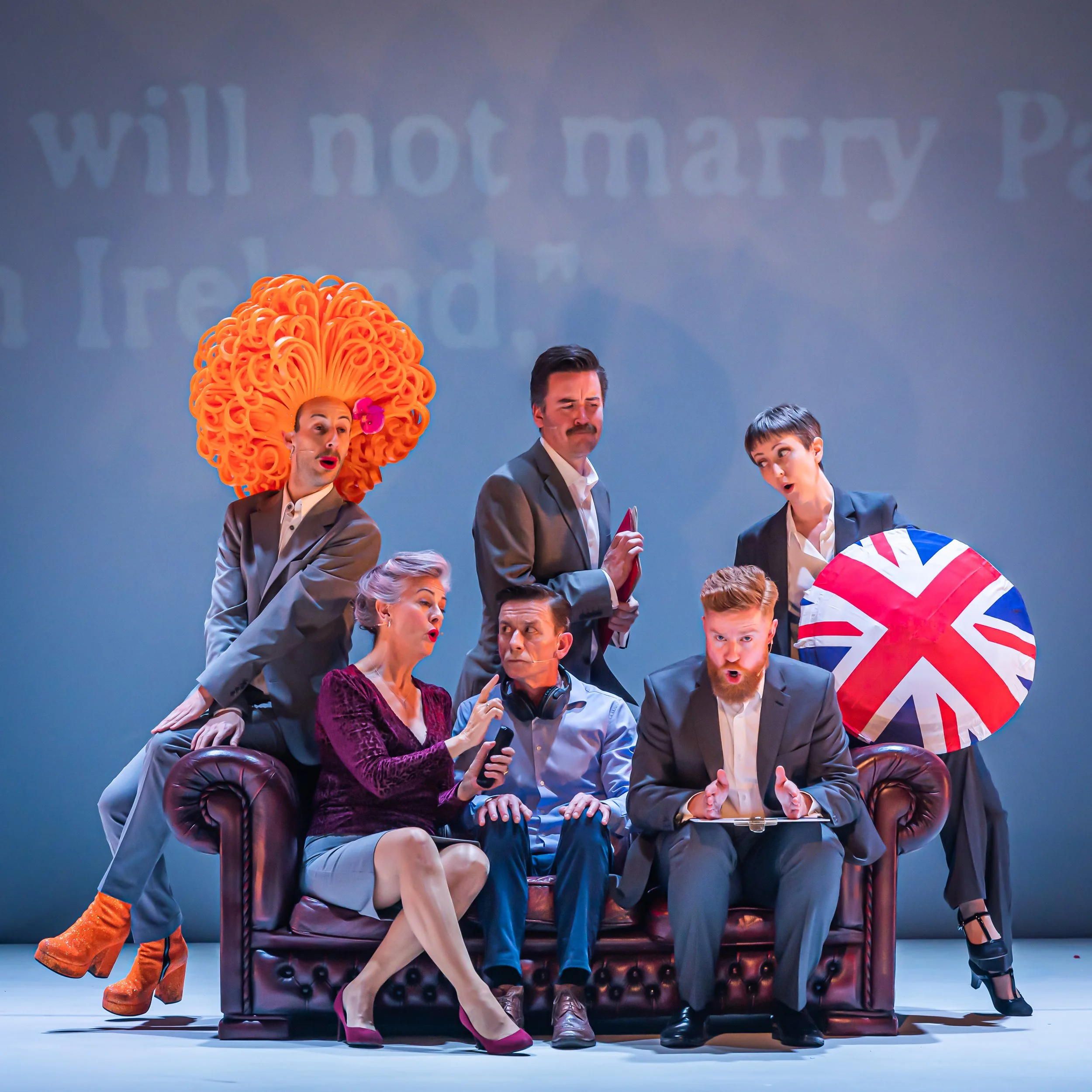 Group of people dressed in formal attire sitting and standing on a stage with a blue background, some making expressive gestures and faces, including a woman with large orange hair and bright pink shoes, a man with a mustache and glasses, and a young