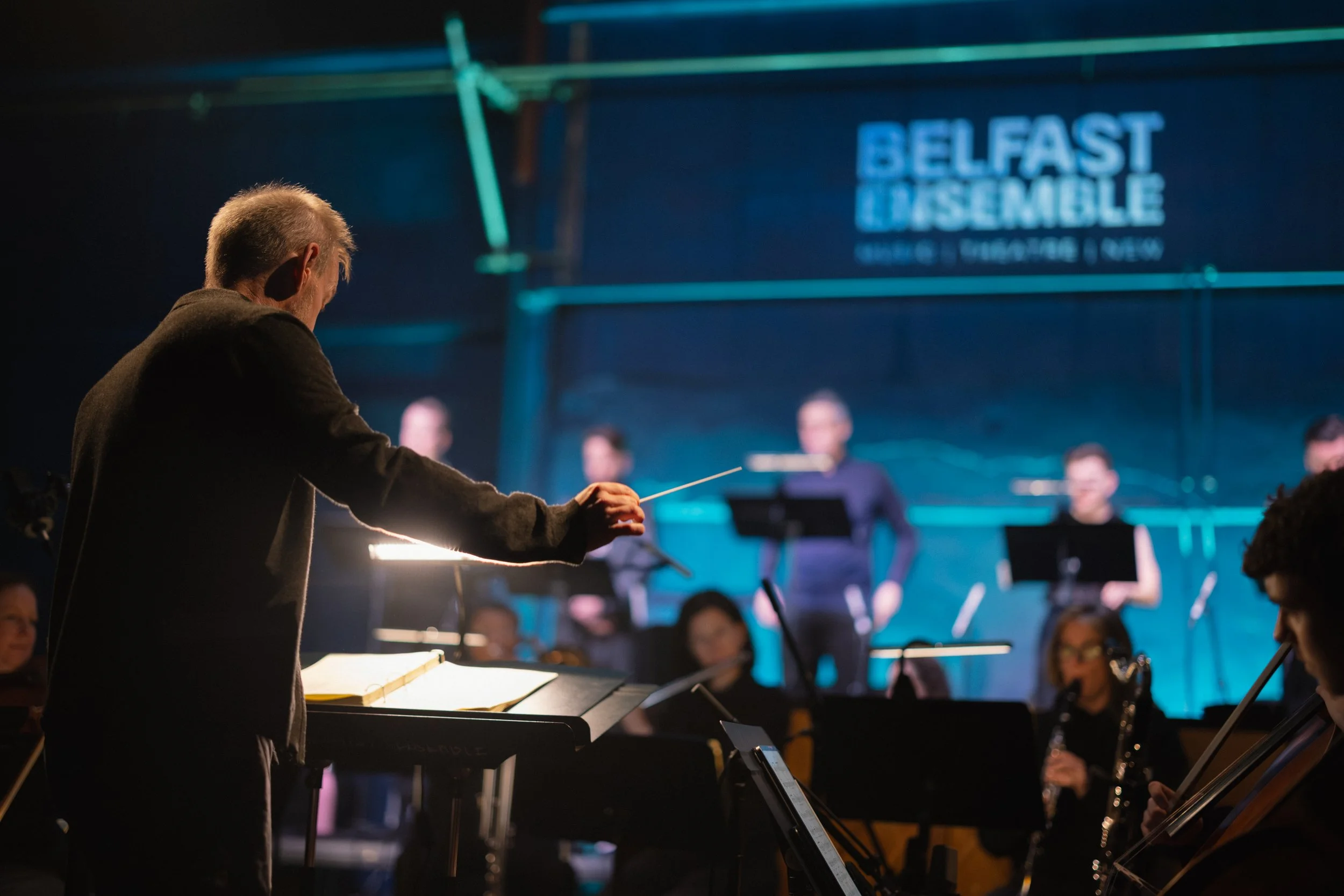 Conductor leading a jazz band on stage at Belfast Limelight, with musicians playing instruments and a blue-lit screen displaying the venue's name.