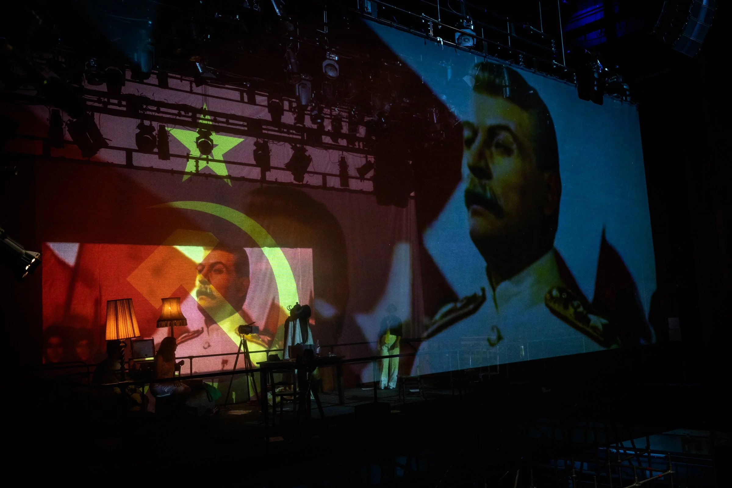 A large projection of a man's face on a screen in a dark, industrial-looking space. The person's face appears serious, and there are stage lights and equipment hanging from the ceiling. The scene is dimly lit with some warm lamp lighting and construc