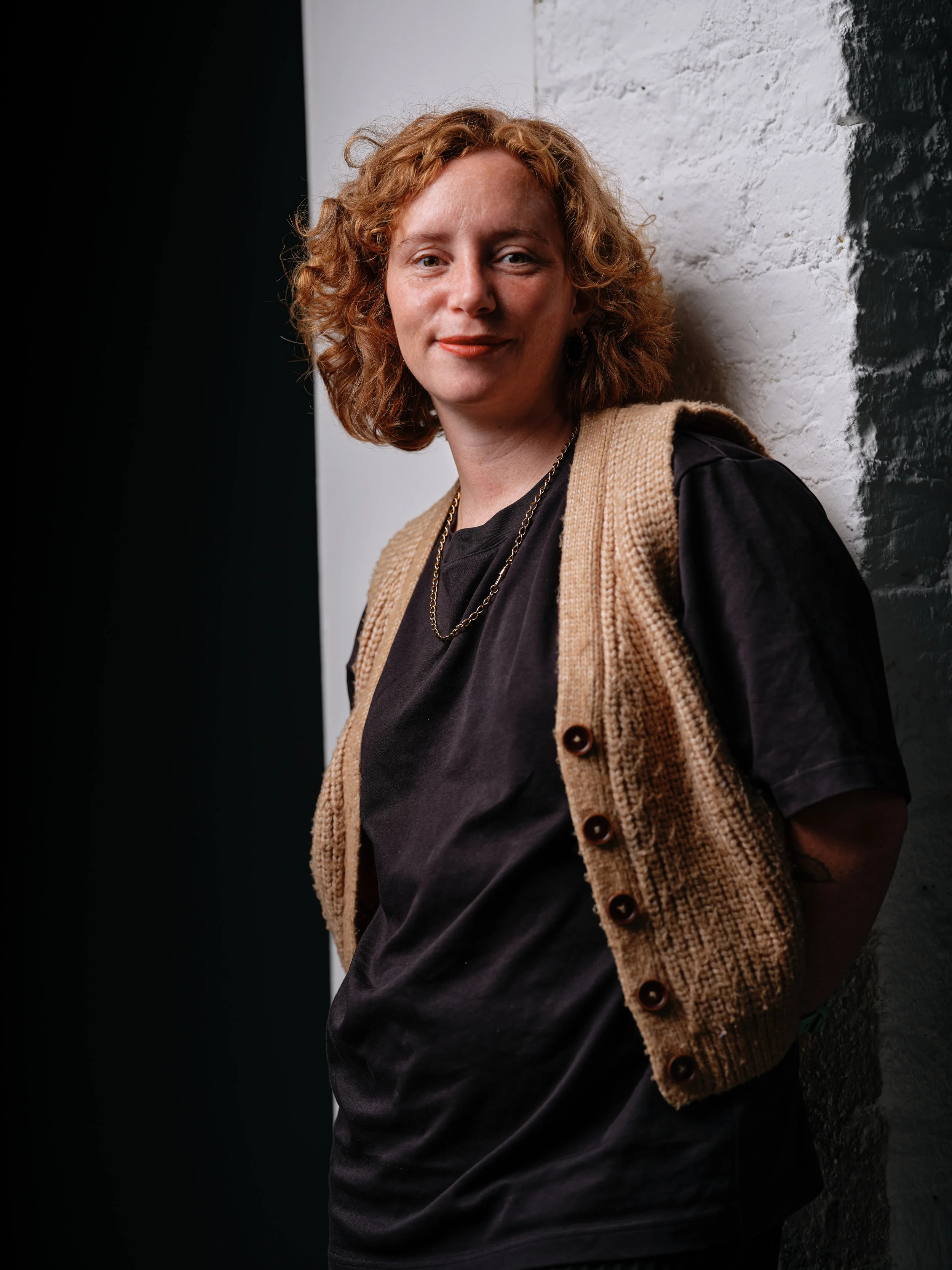 A woman with curly red hair, wearing a black shirt, a tan knitted vest, and gold jewelry, leaning against a white and black textured wall.