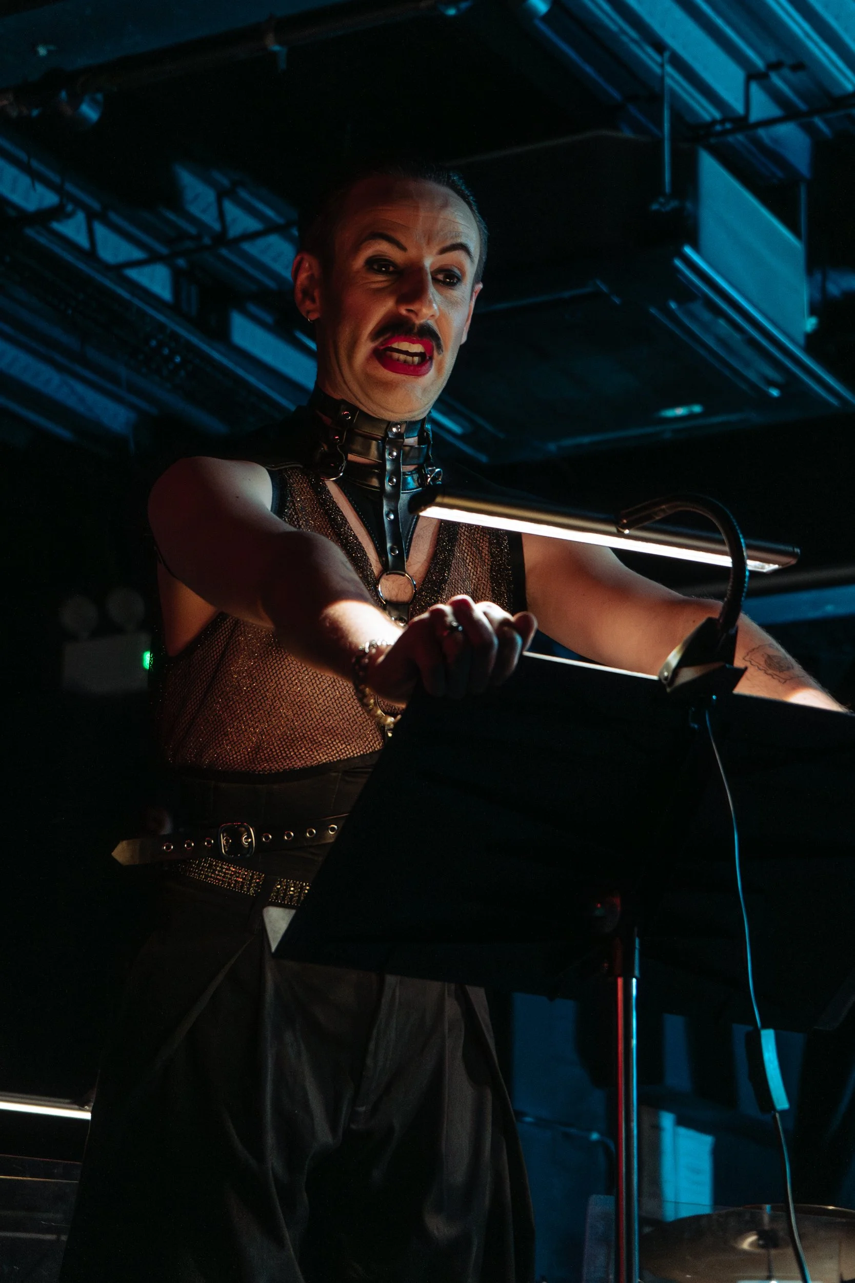 A person with a slicked-back hairstyle, makeup, and a leather outfit, performing at a nightclub or bar, illuminated by dim lighting.