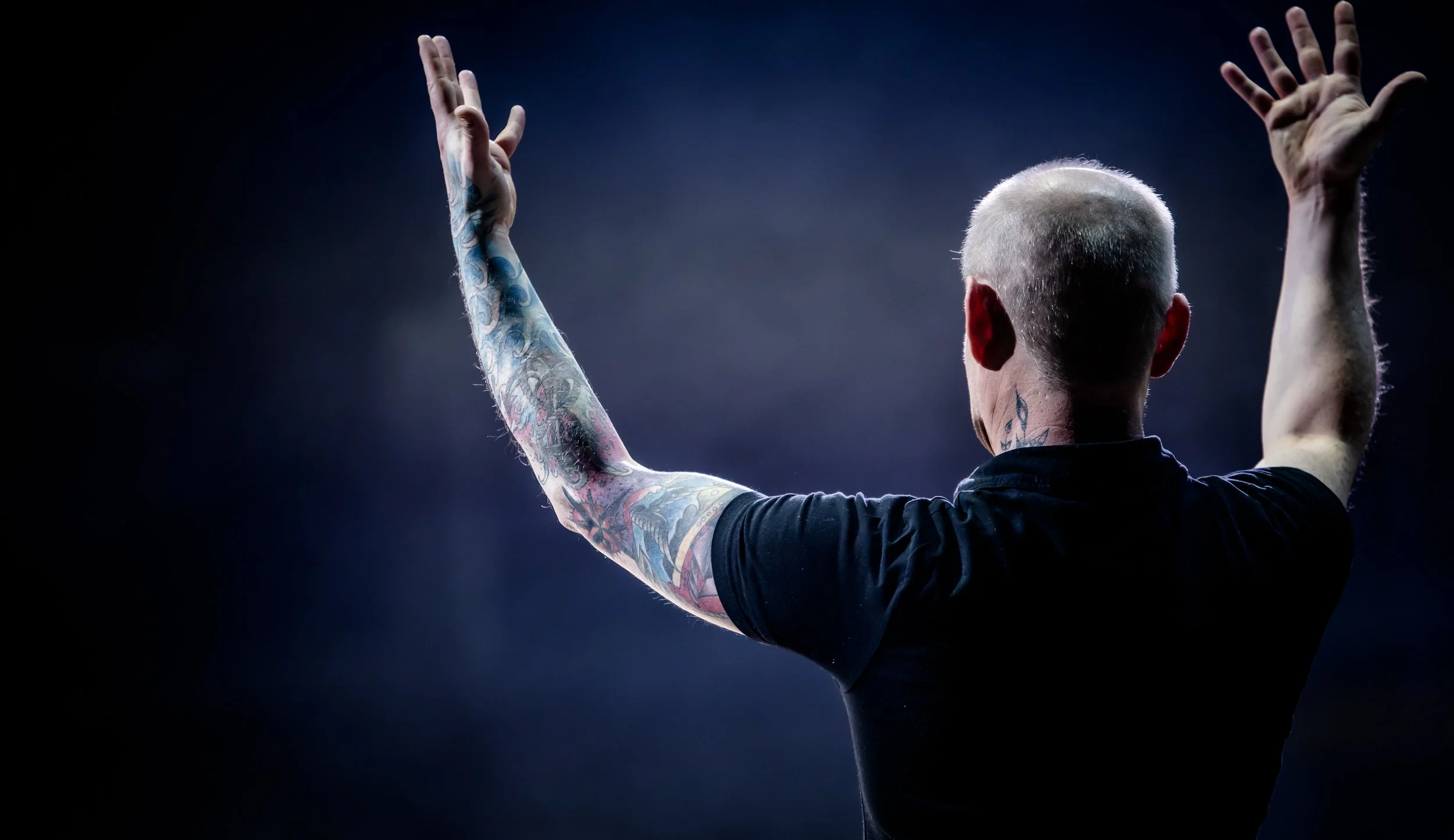 A person with tattoos on their left arm and back, with short, gray hair, raising both arms, viewed from behind, against a dark background.