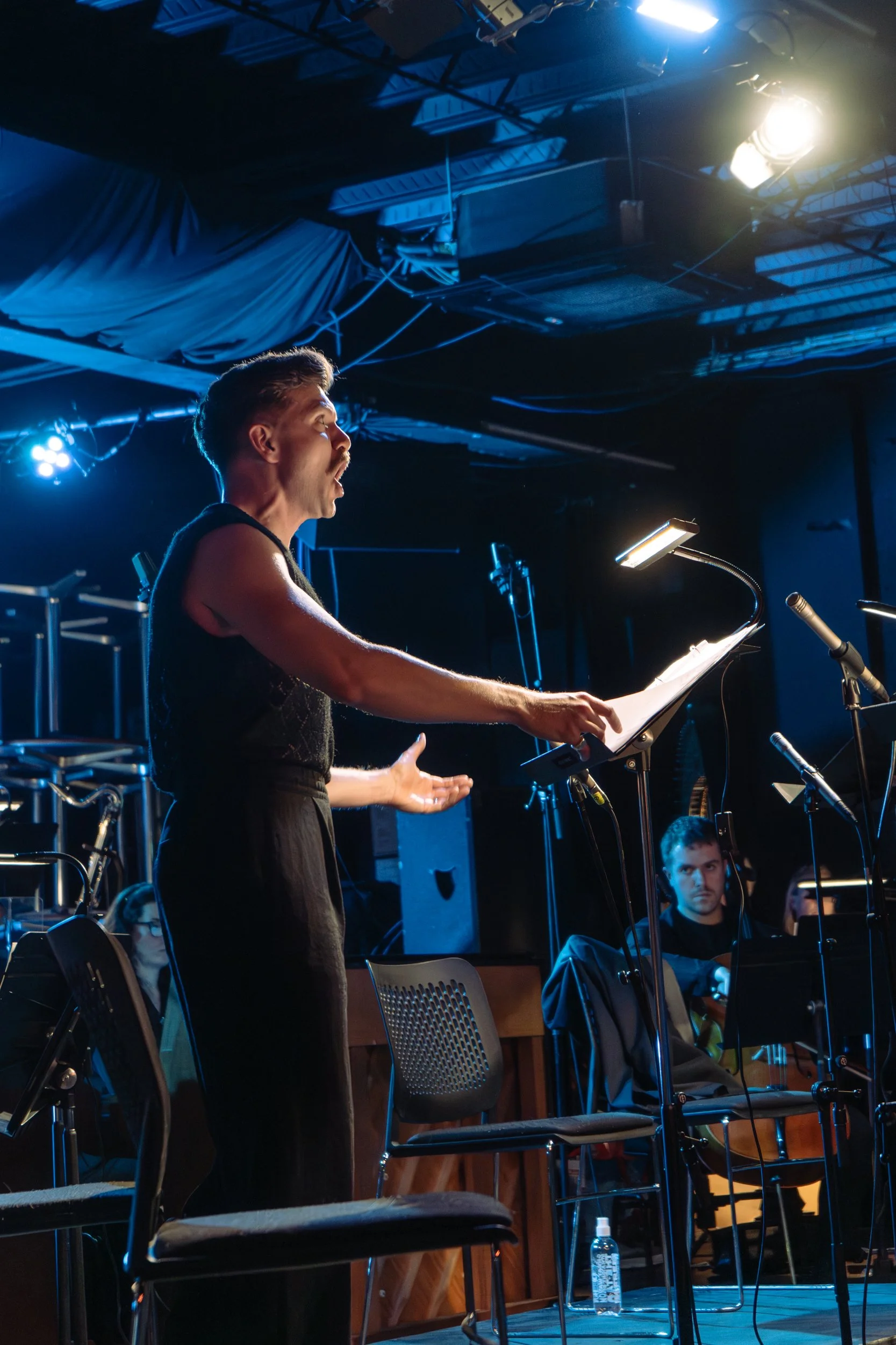 A woman with short hair and a mustache, wearing a sleeveless black top, is standing on stage during a performance or rehearsal, singing or speaking with her mouth open and hands gesturing. In the background, there are seated musicians and music stand