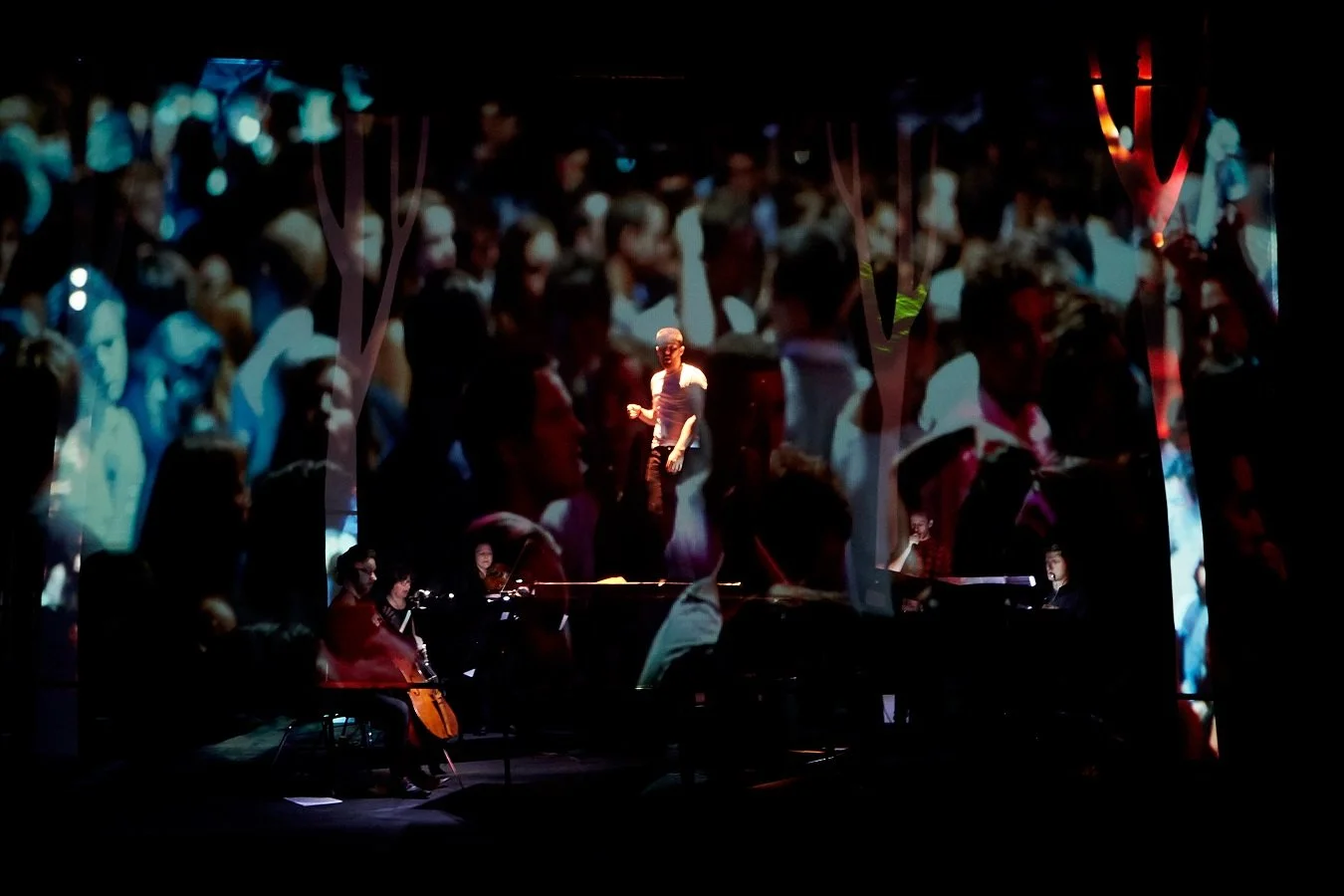 A live performance with a woman singing on stage, surrounded by a multimedia projection of a crowd of people, and accompanied by musicians playing piano and cello.