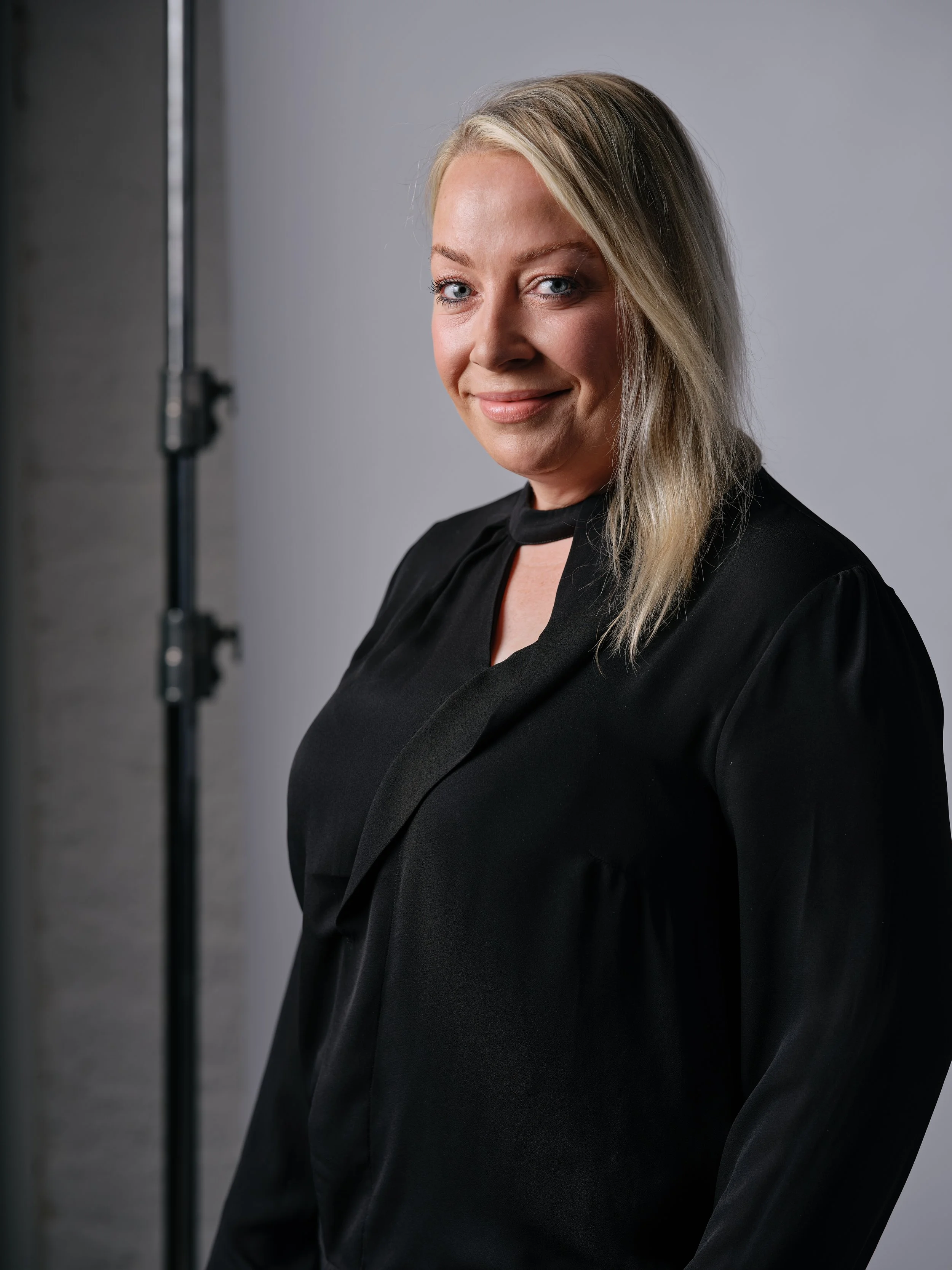 A woman with blonde hair and blue eyes smiling, wearing a black outfit, standing in front of a plain gray background.