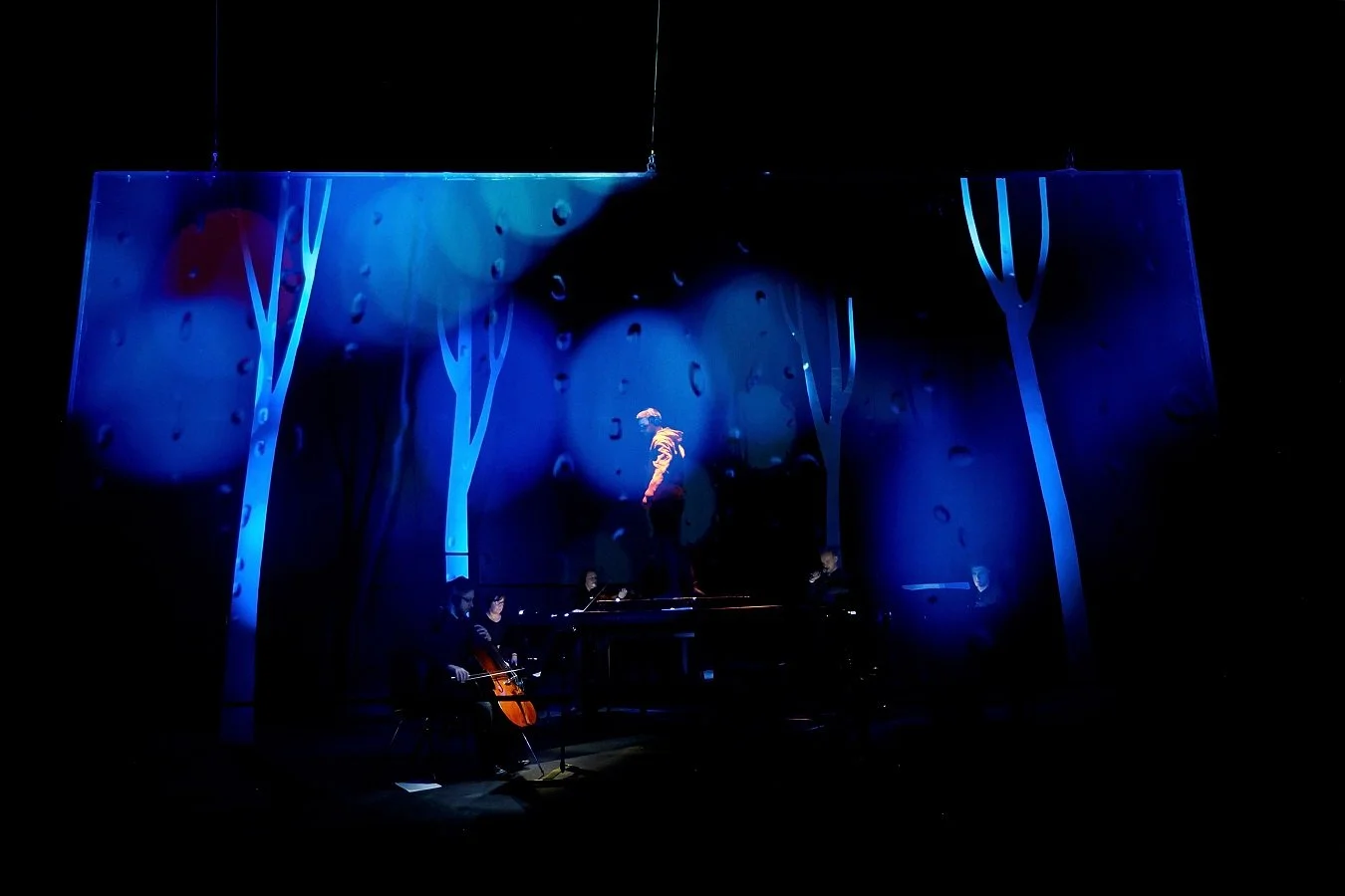 A theater stage with a small orchestra and a performer in the center, surrounded by blue-lit tree cutouts and a cloudy background with raindrop visuals.
