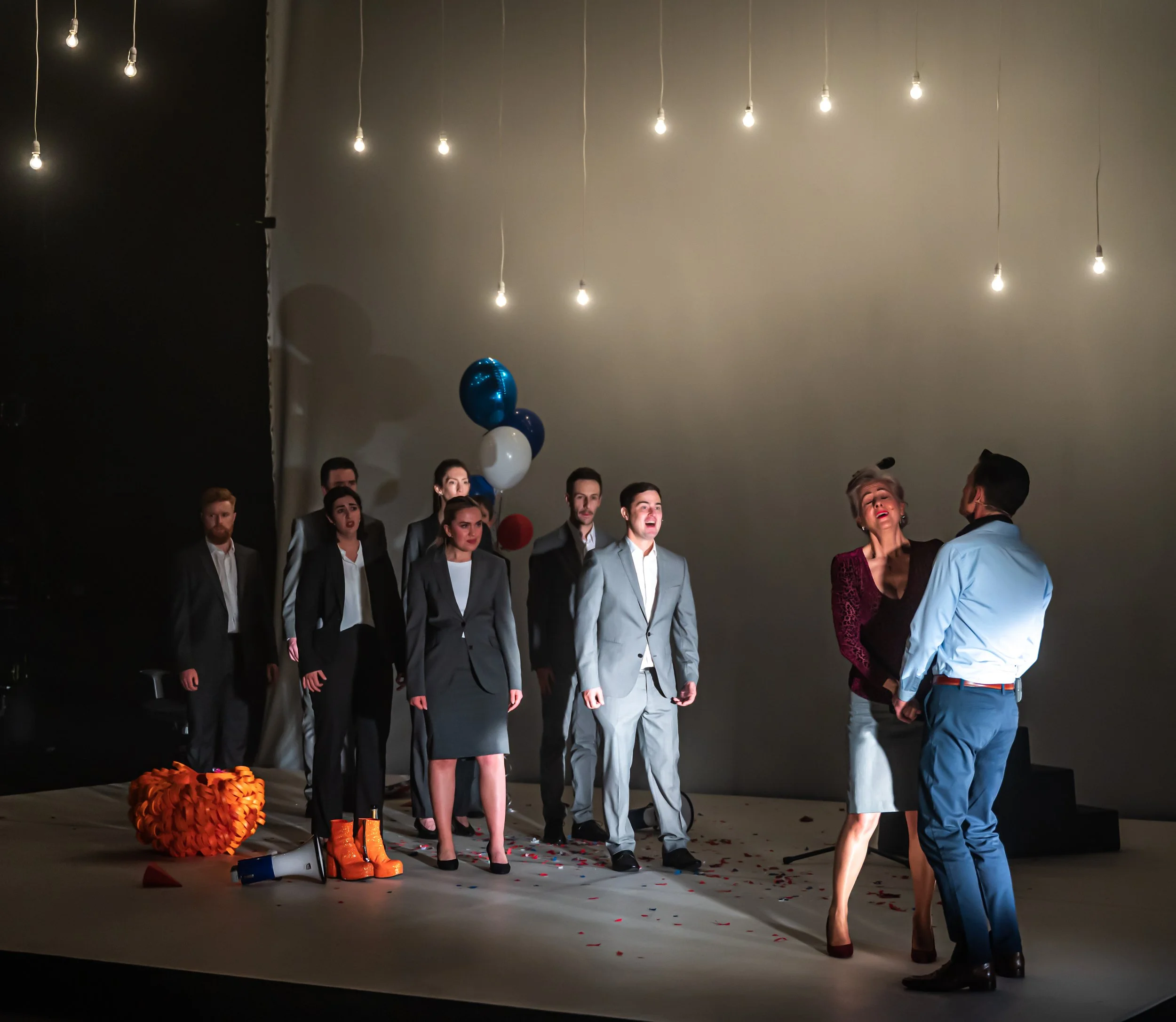 A group of people in formal attire standing on a stage with a plain wall background, decorated with blue and white balloons, orange shoes, and orange decorations on the floor, with hanging lights overhead.
