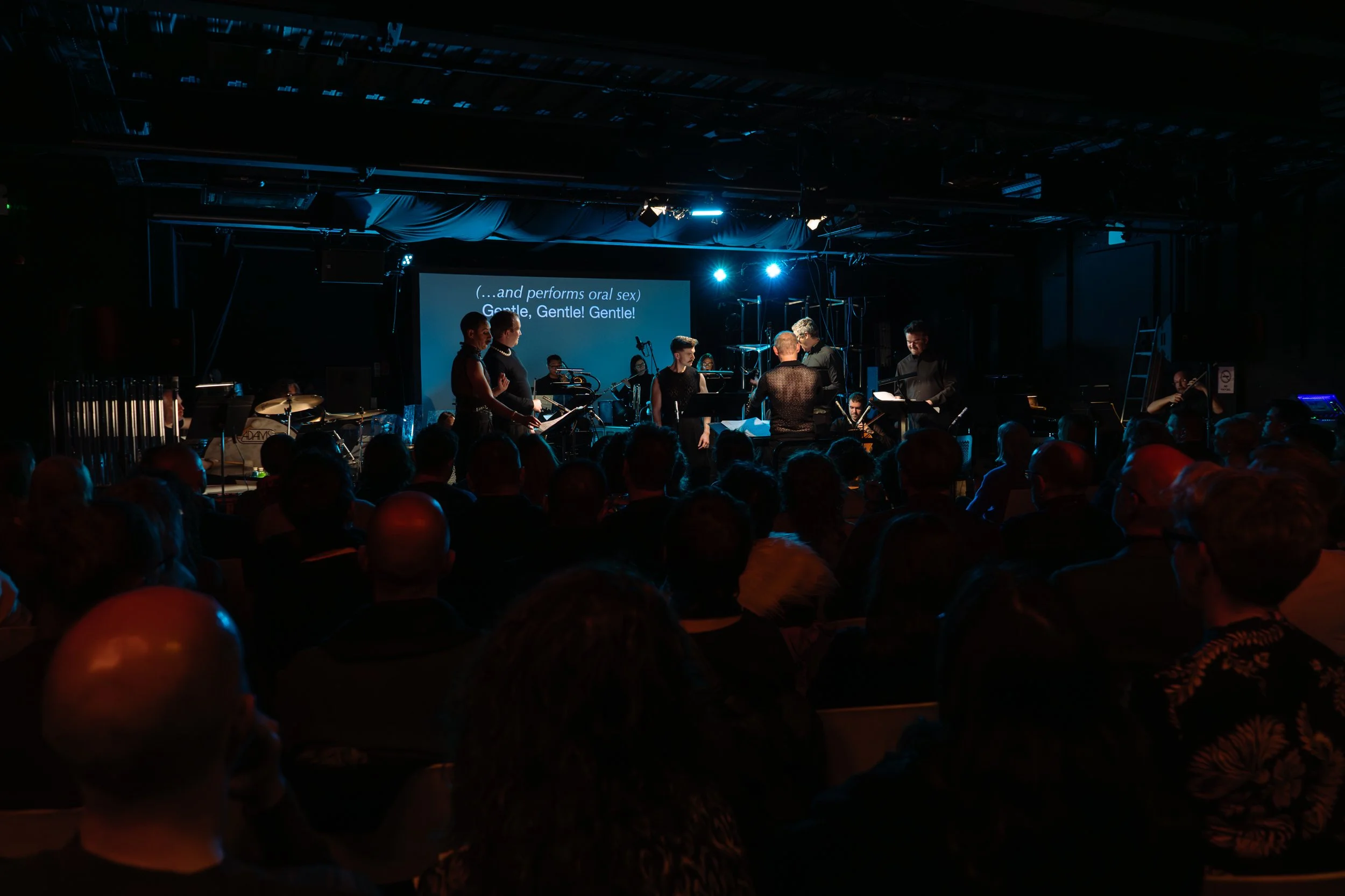 A live performance on stage with musicians and singers, audience in foreground, large screen displaying lyrics, dark lighting with blue spotlights.