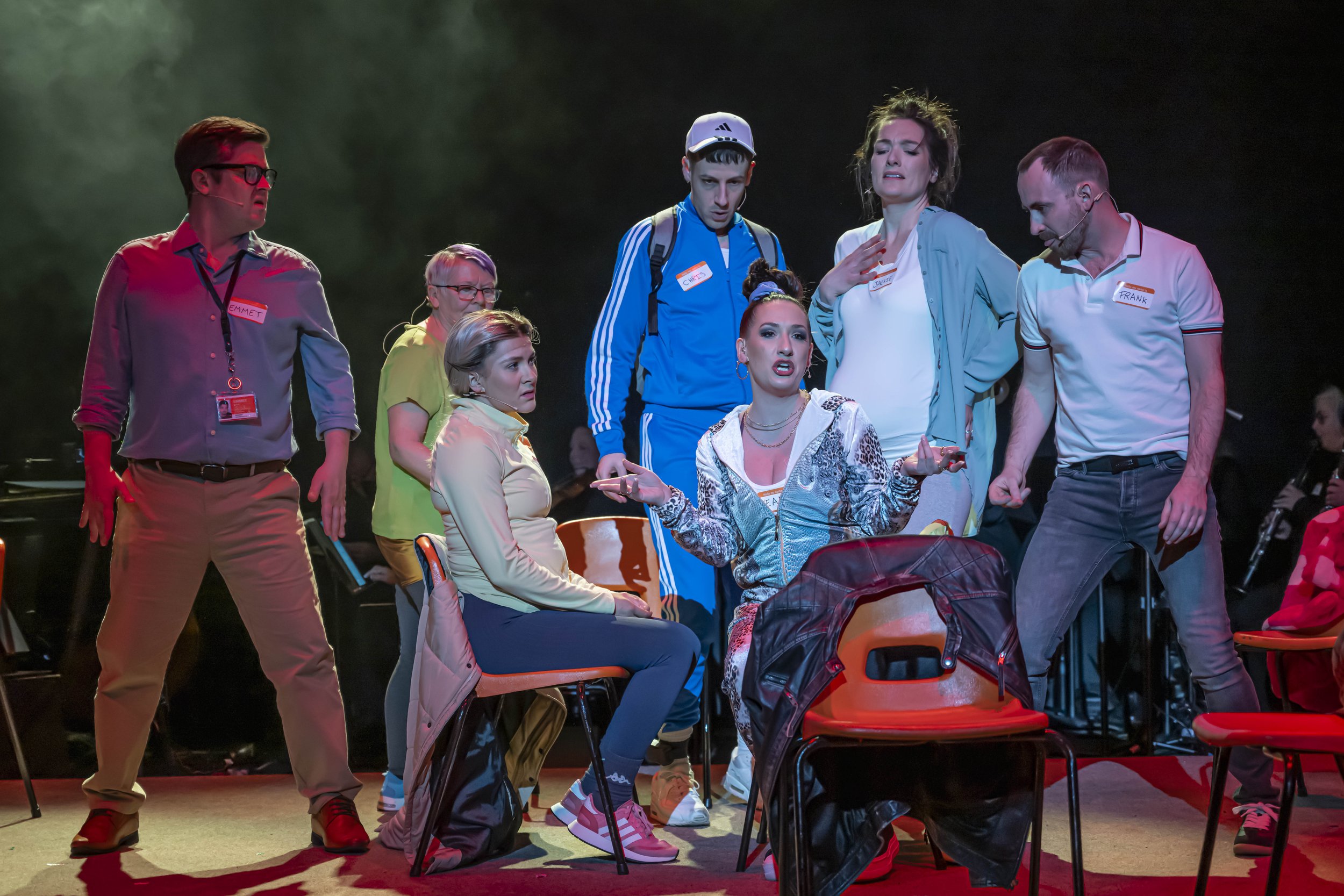 A group of actors on stage in a dramatic scene with serious expressions, seated and standing around a table, some wearing colorful costumes and name tags.
