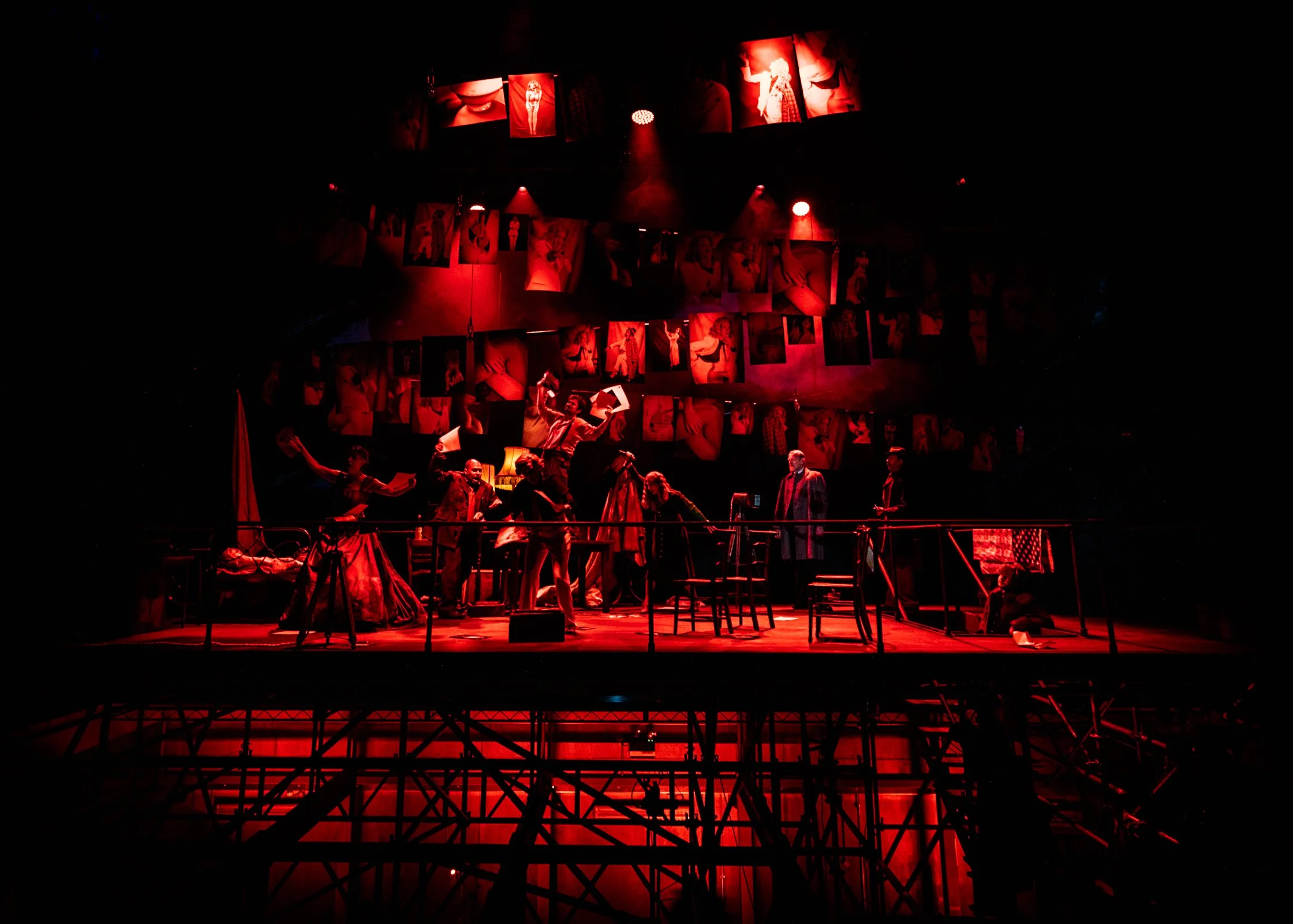 Stage scene with red lighting, multiple actors in costumes, photographs hanging from ceiling, and a raised platform with chairs.