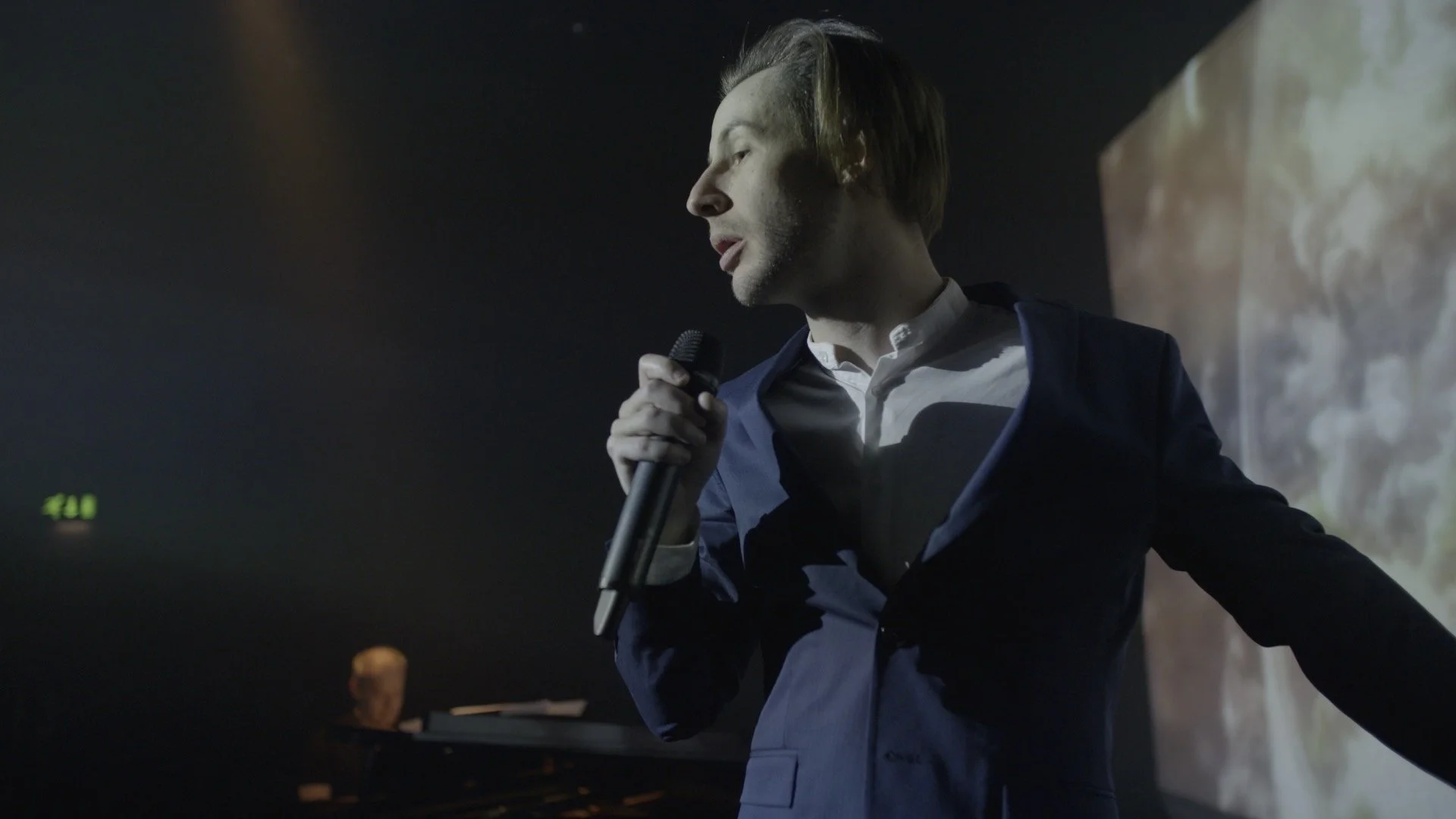 A man in a dark suit and white shirt holding a microphone while looking to his right, standing near a large screen or wall made of stone or concrete.
