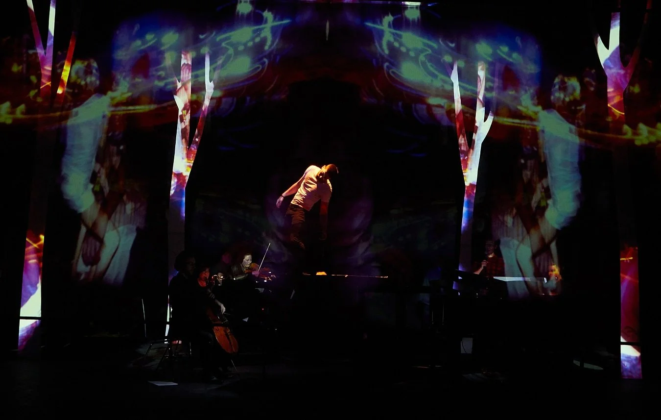 A multimedia performance with a person jumping in the air against a backdrop of colorful, abstract digital projections, with musicians playing instruments in front.