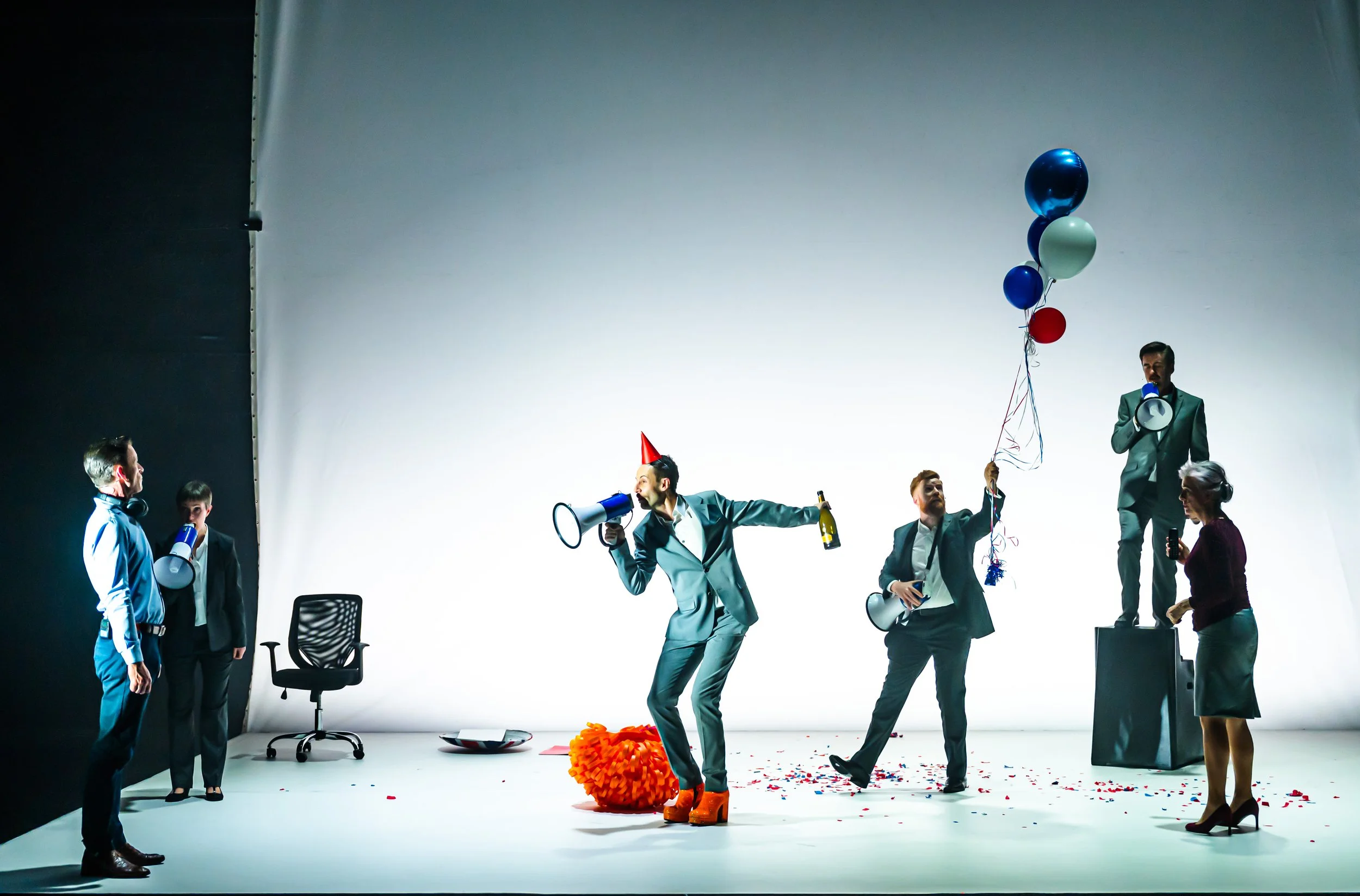 Group of people celebrating in a photo studio with balloons, megaphones, and confetti, some wearing party hats and costumes.