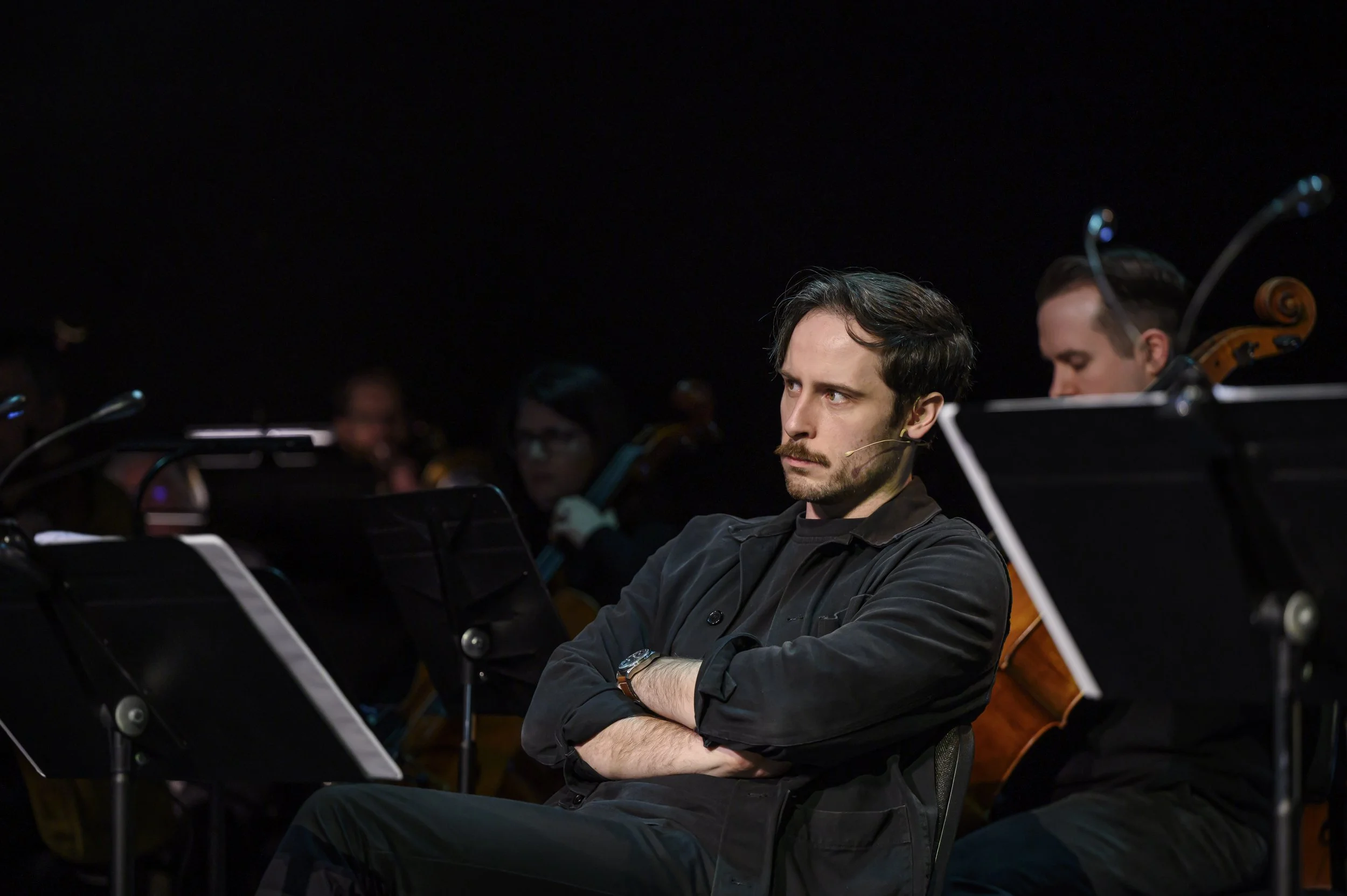 A man with dark hair, a beard, and a serious expression, sitting with arms crossed, in a dark setting among musicians with sheet music stands and music instruments.