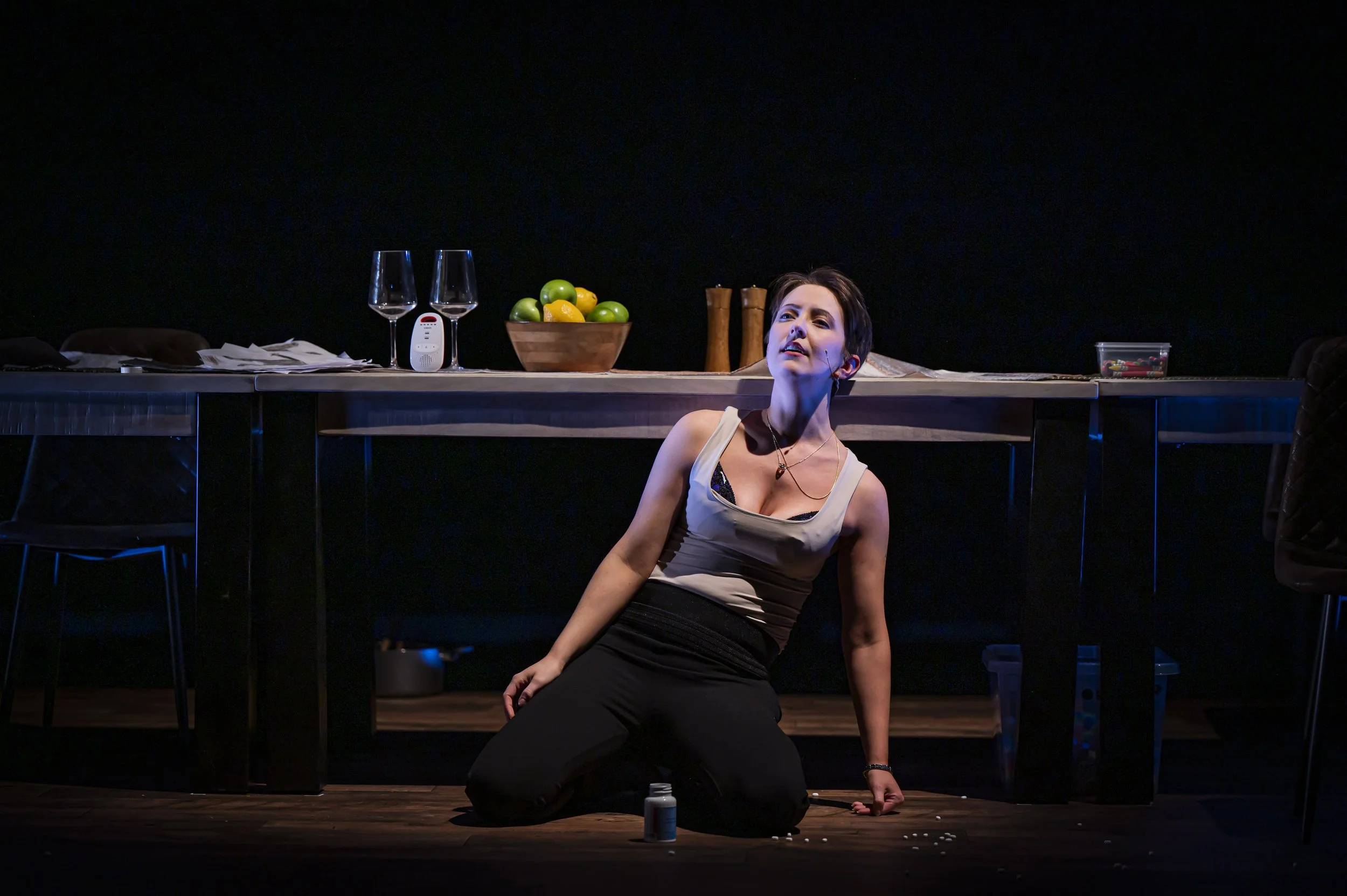 A woman is performing on stage, kneeling on the floor with her head tilted back and one arm resting on her knee. A table behind her holds two empty wine glasses, a bowl of green apples and lemons, salt and pepper shakers, a small white device, and a 