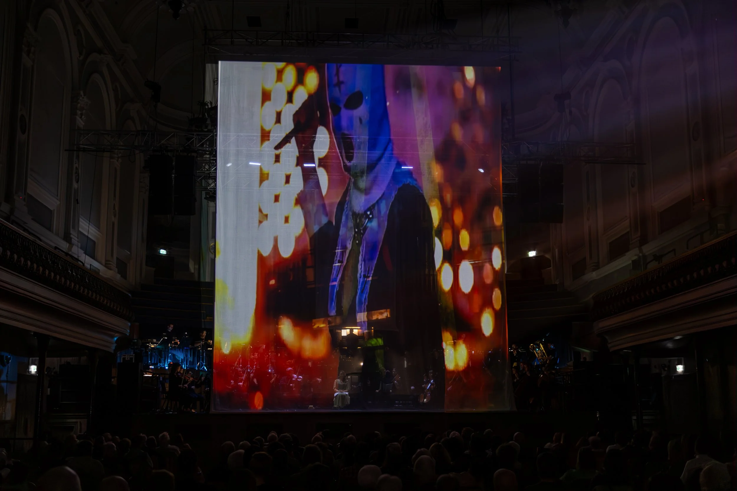 Large indoor venue with an expansive digital screen displaying abstract colorful visuals, with an audience seated below observing the show.