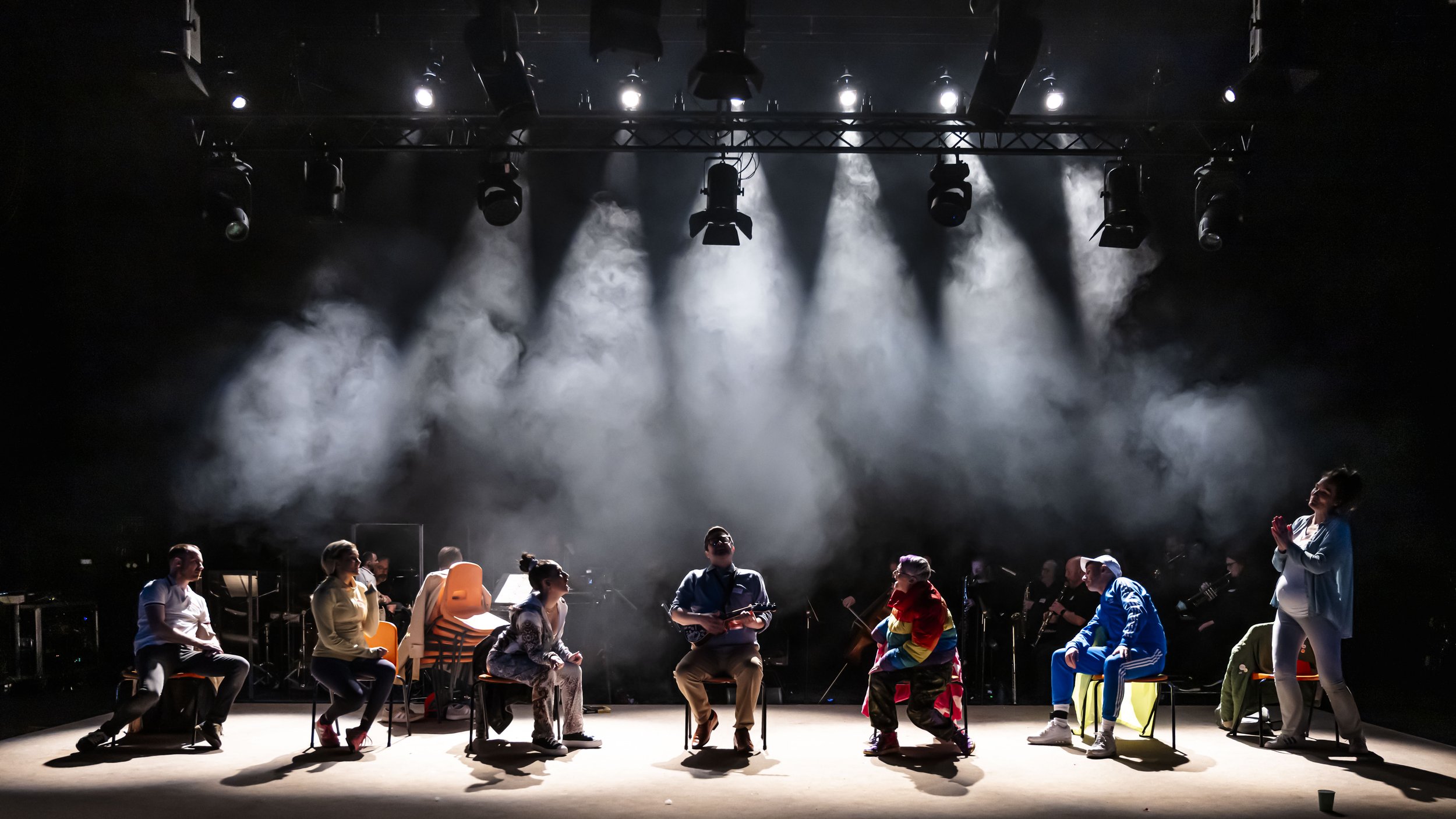 A group of people sitting and standing on a stage with stage lights and fog, possibly rehearsing or performing.