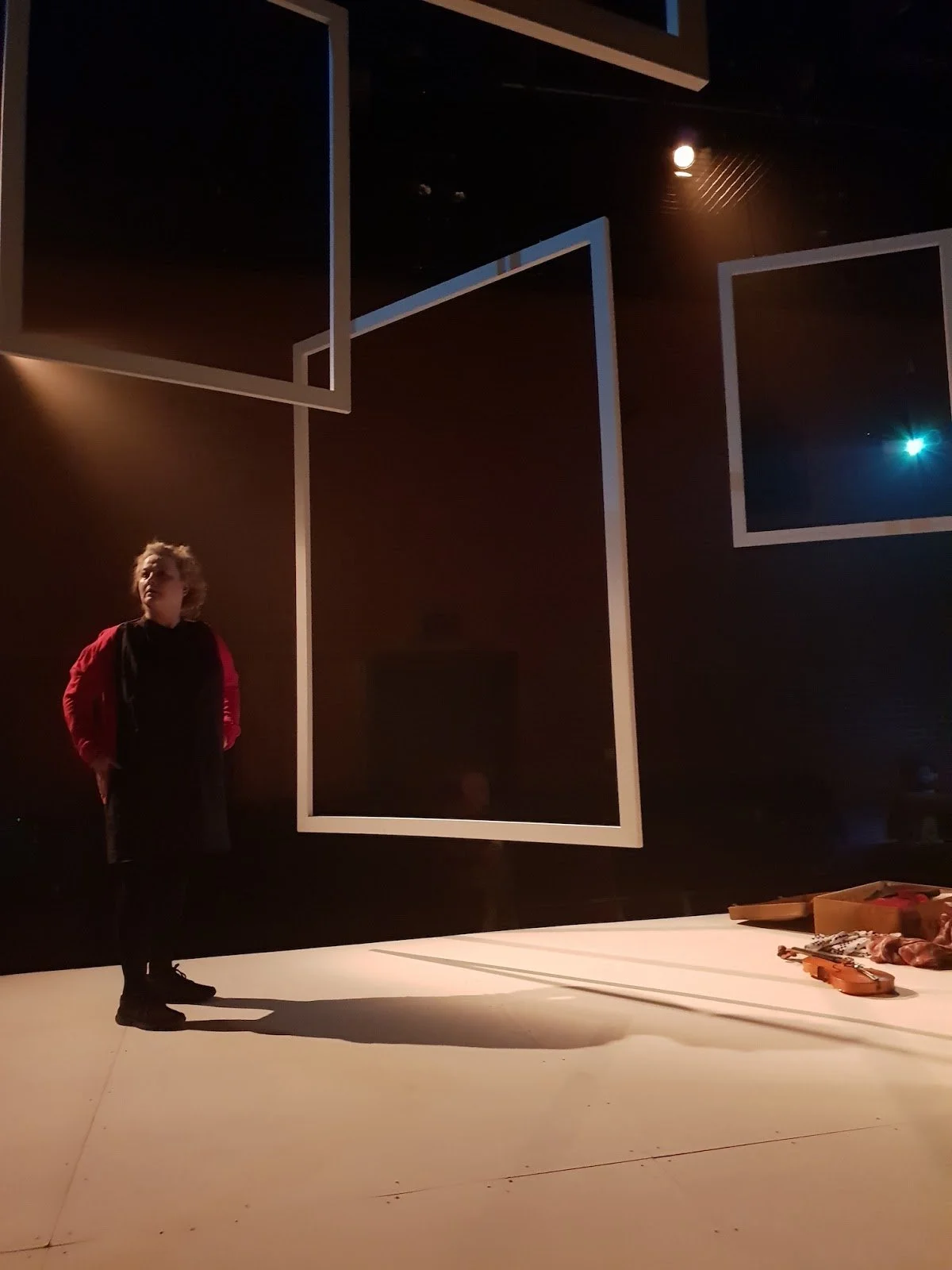 A woman stands on a stage with a spotlight on her, surrounded by large empty picture frames hanging from the ceiling. There is a musical instrument and a box on the stage floor.