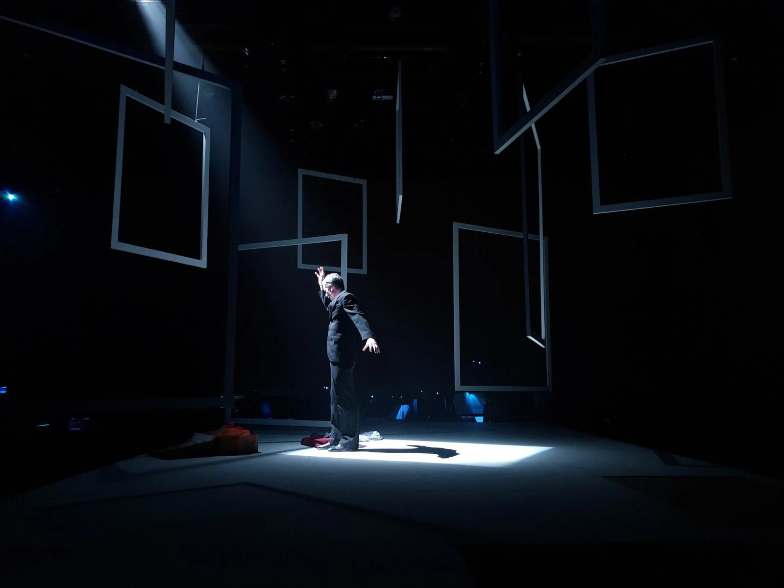 A person standing on a stage with a spotlight, surrounded by hanging empty picture frames in a dark setting.