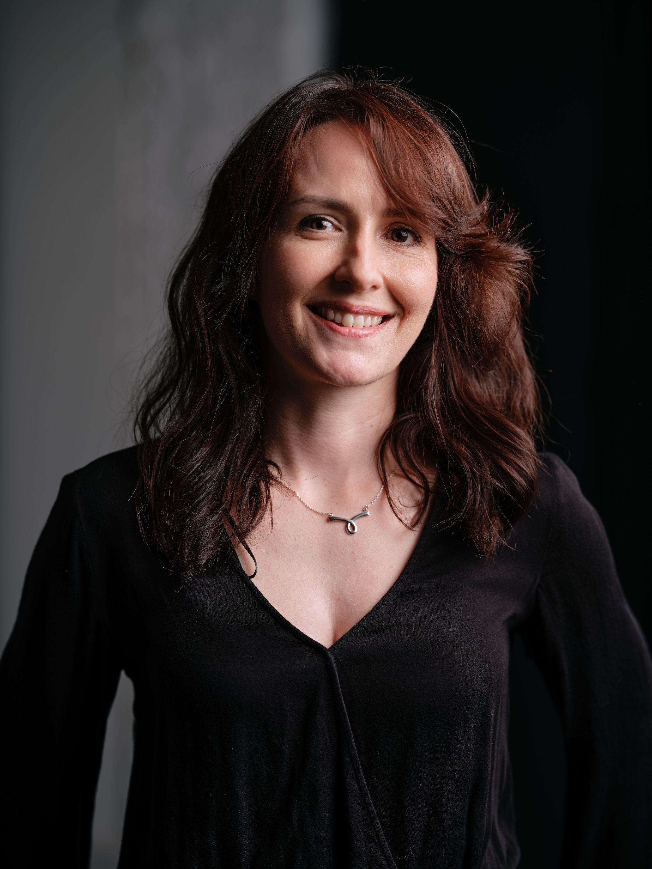 Close-up of a woman with shoulder-length wavy brown hair, smiling and wearing a black top and a necklace with a bird-shaped pendant, against a dark background.
