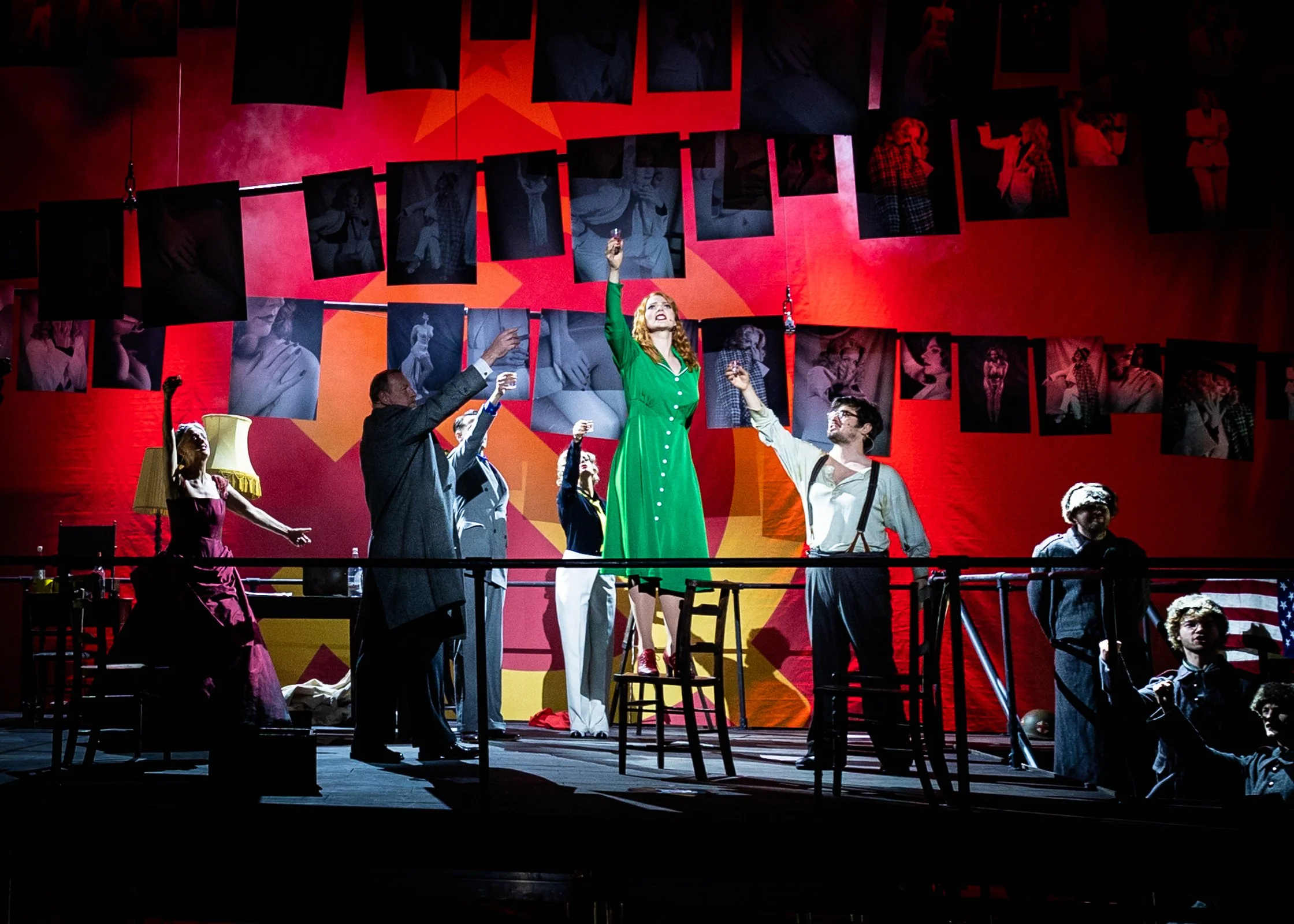 Theatrical performance on stage with a woman in a green dress holding a drink and raising her arm, surrounded by people, with photographs or posters hanging on a red and orange background.