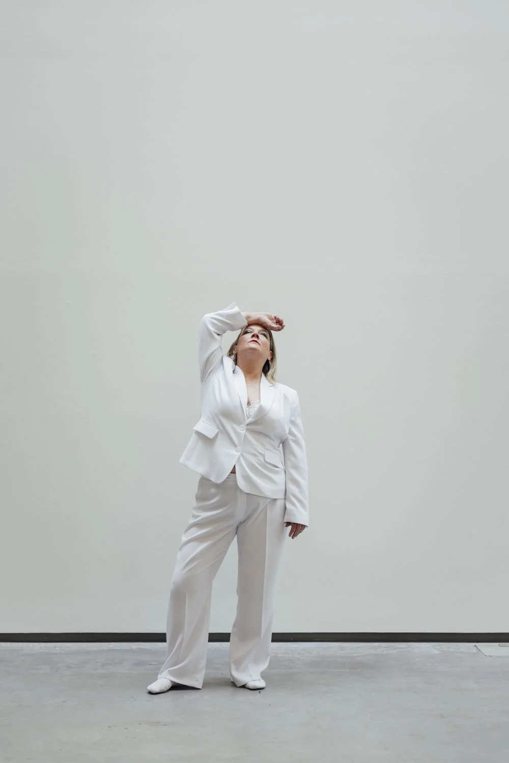 A woman dressed in a white business suit standing in front of a plain light-colored wall, with her hand on her forehead looking upward.