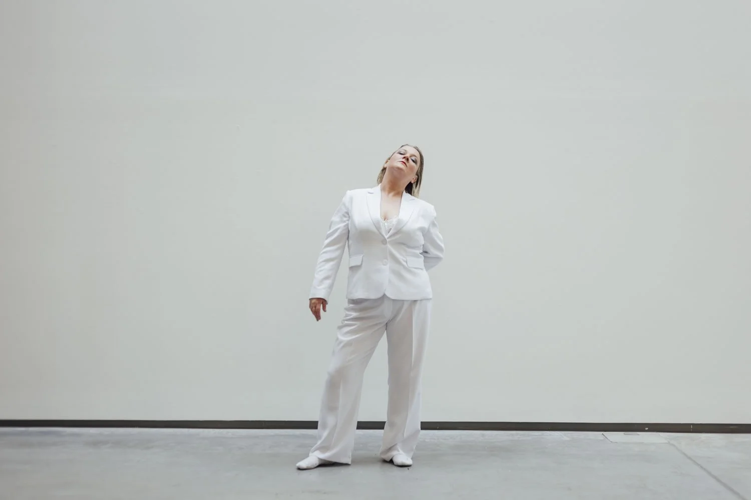 A woman in a white suit standing against a plain light gray wall, leaning her head back with eyes closed.