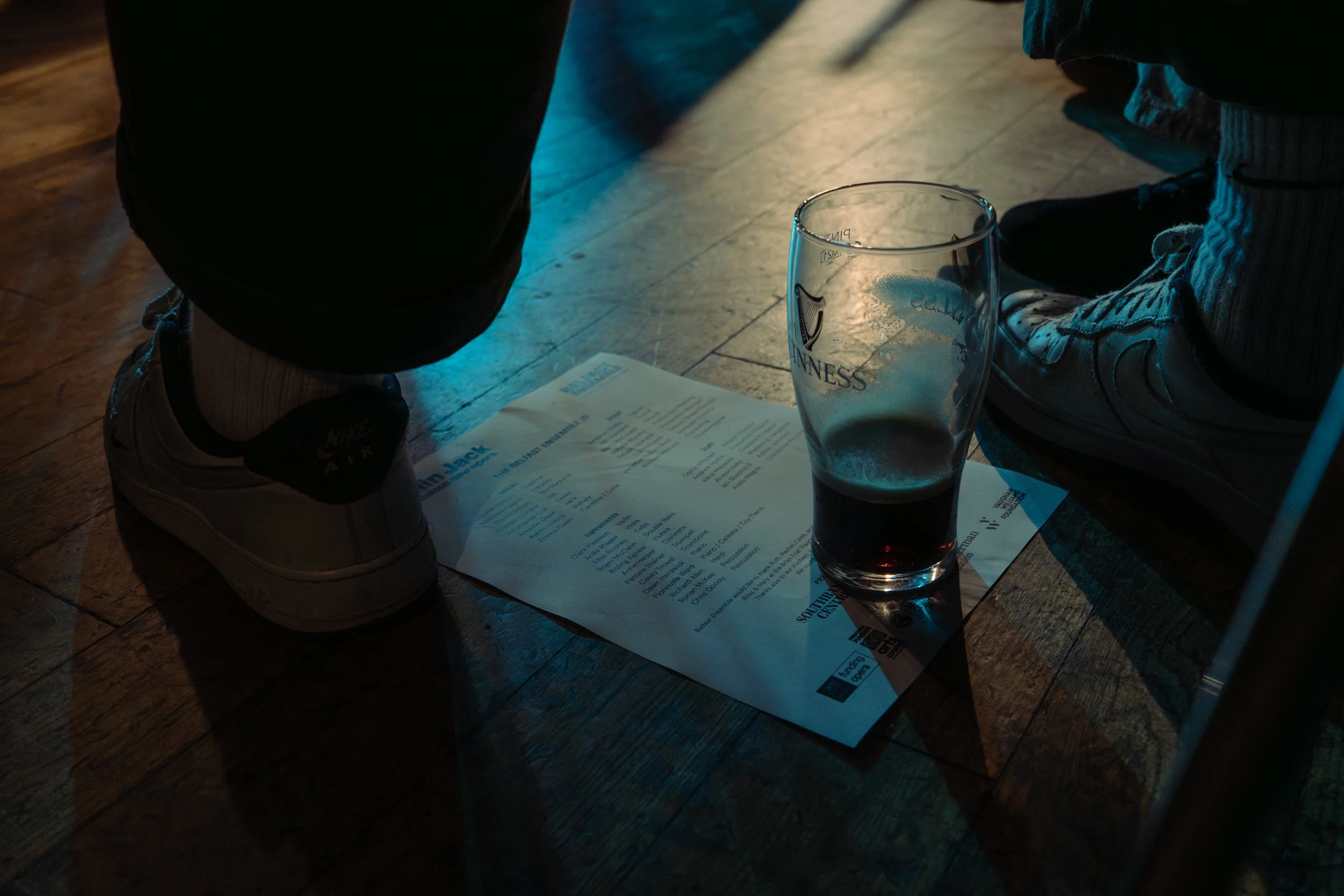 Close-up of a glass of Guinness beer, a beer menu on the floor, and part of a person's legs and shoes, taken indoors with dim lighting.