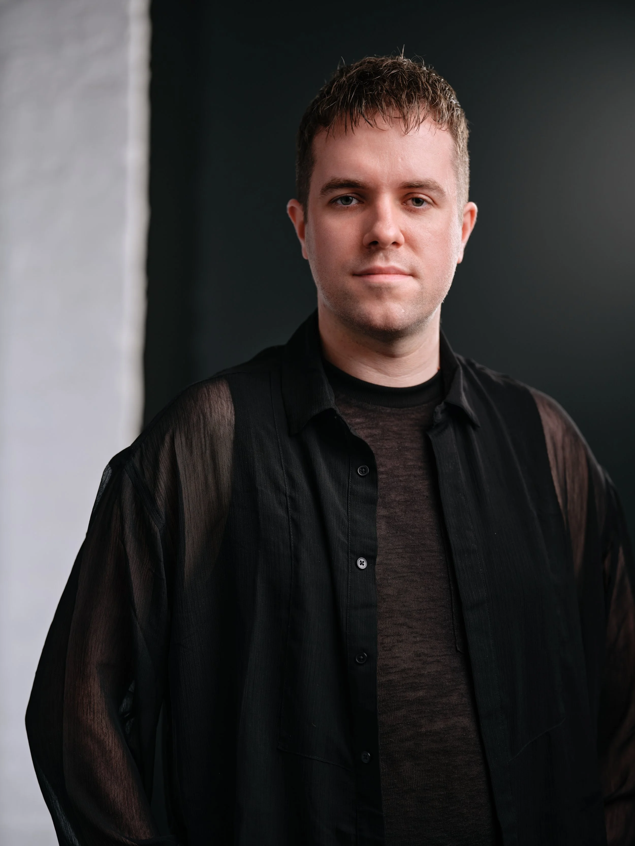 Portrait of a young man with short brown hair, wearing a black shirt, standing against a dark background with a white wall on the left side.