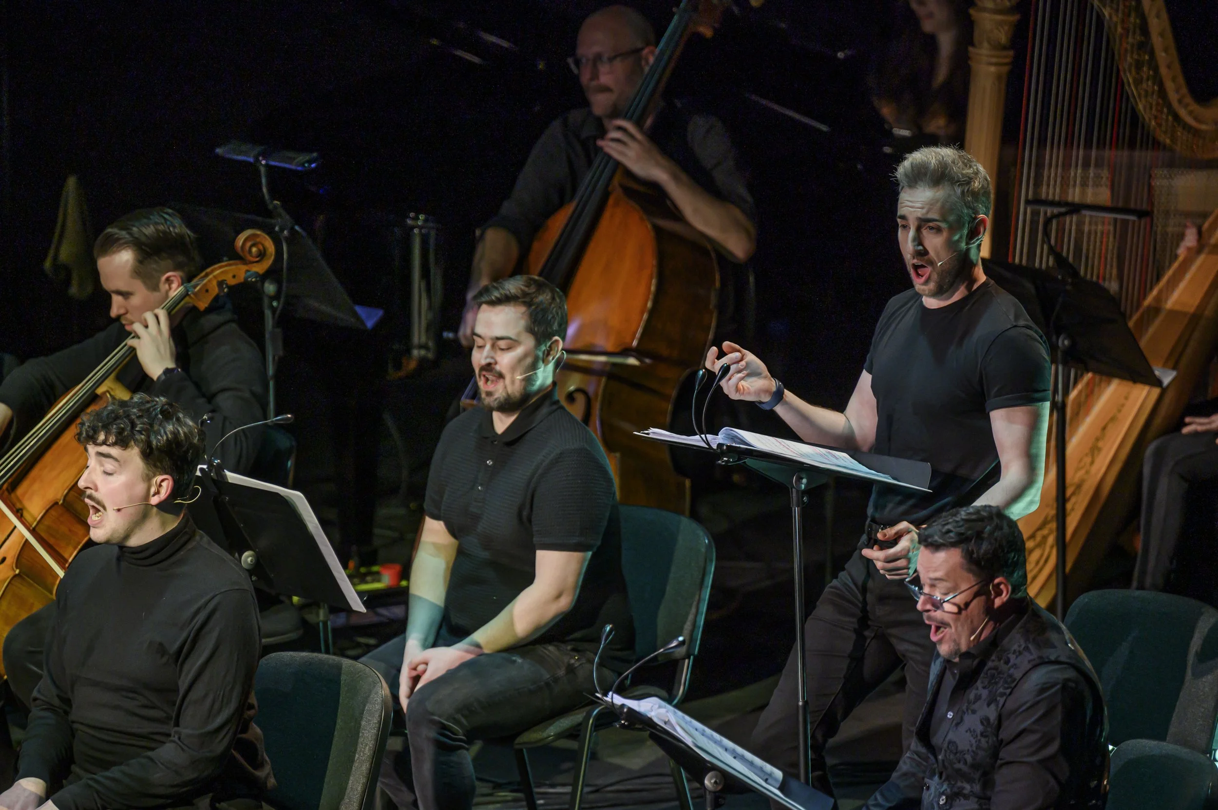 Musicians performing on stage, singing and playing instruments including a cello, harp, and other string instruments, with focused and passionate expressions.