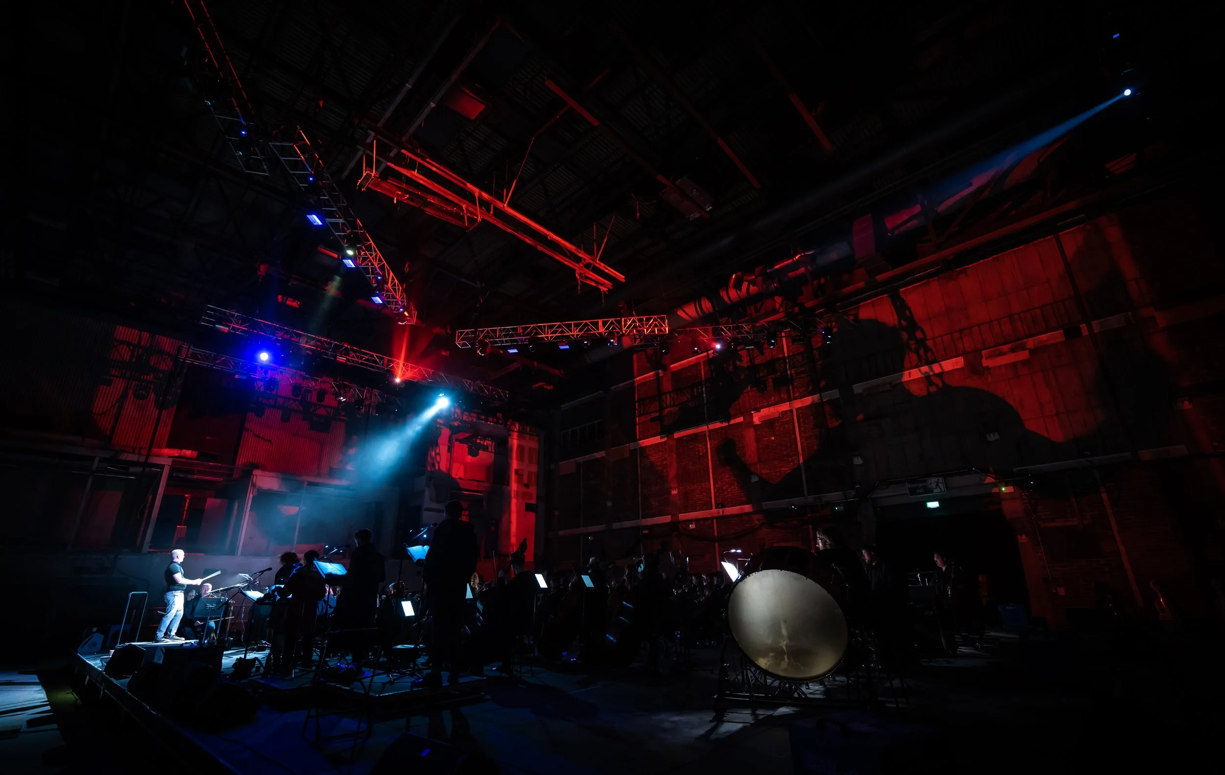 An indoor stage with musicians and orchestral instruments under red and blue lighting, with scaffolding and structural elements on the ceiling.