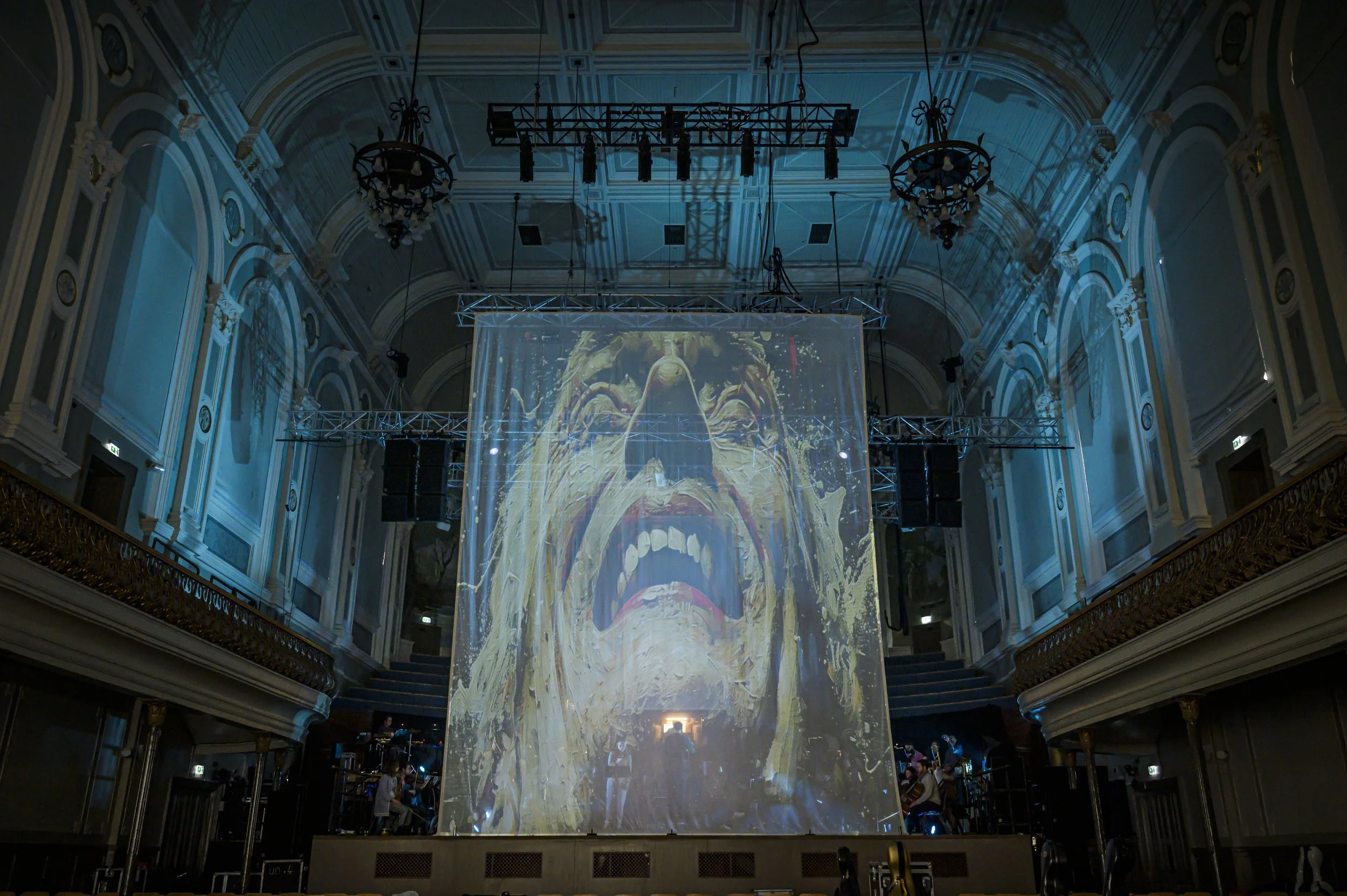 An ornate hall with high, decorated ceilings and chandeliers, featuring a large projection screen displaying an image of a screaming face.