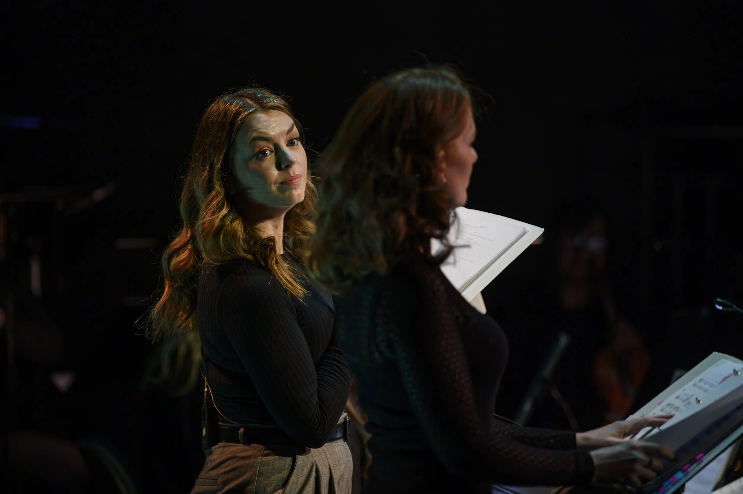 Two women, one with red hair and the other with dark hair, are standing in a dimly lit room, reading from scripts or music sheets.