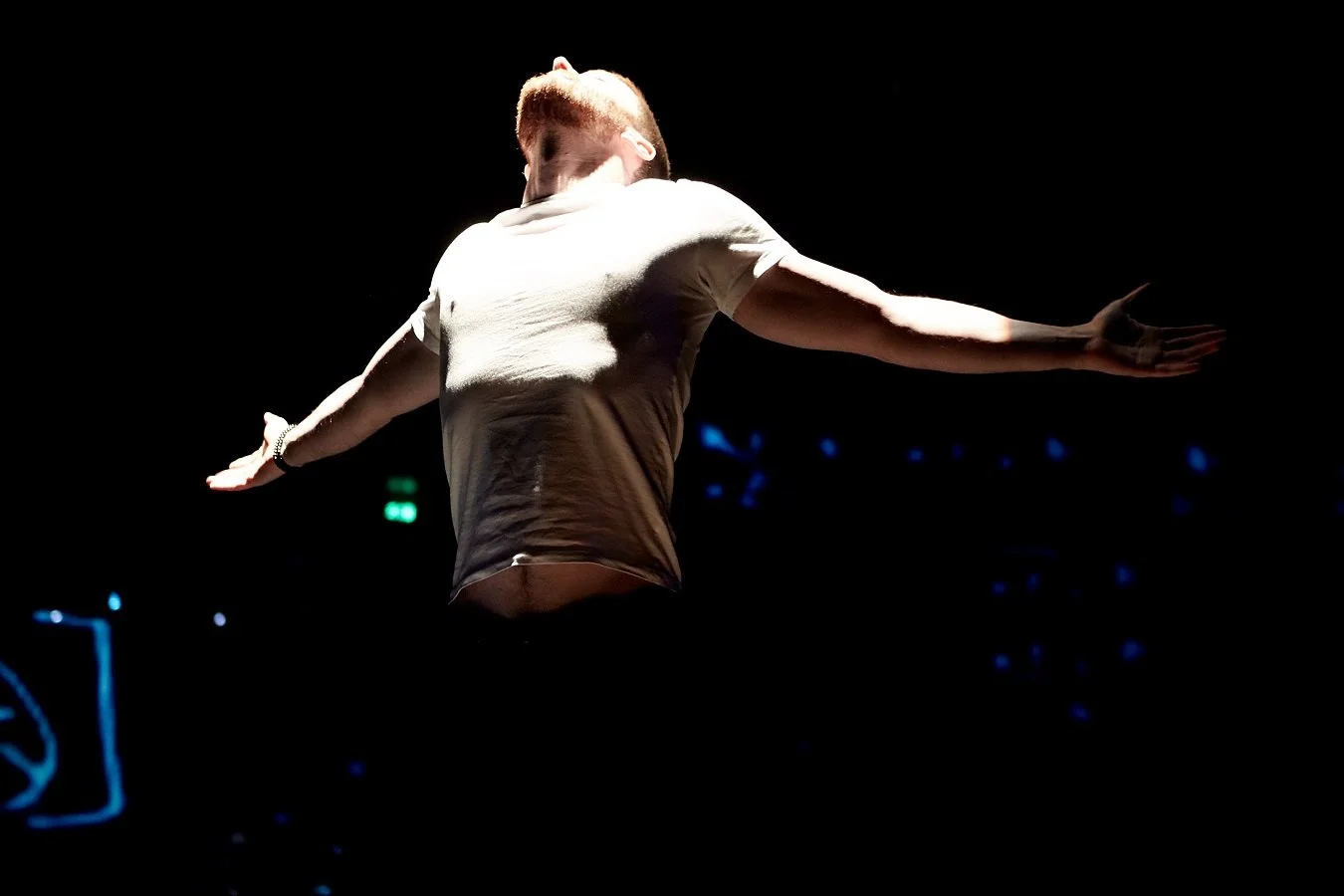 A person with red hair wearing a white shirt with arms extended, standing in a dark environment with bright stage lighting.