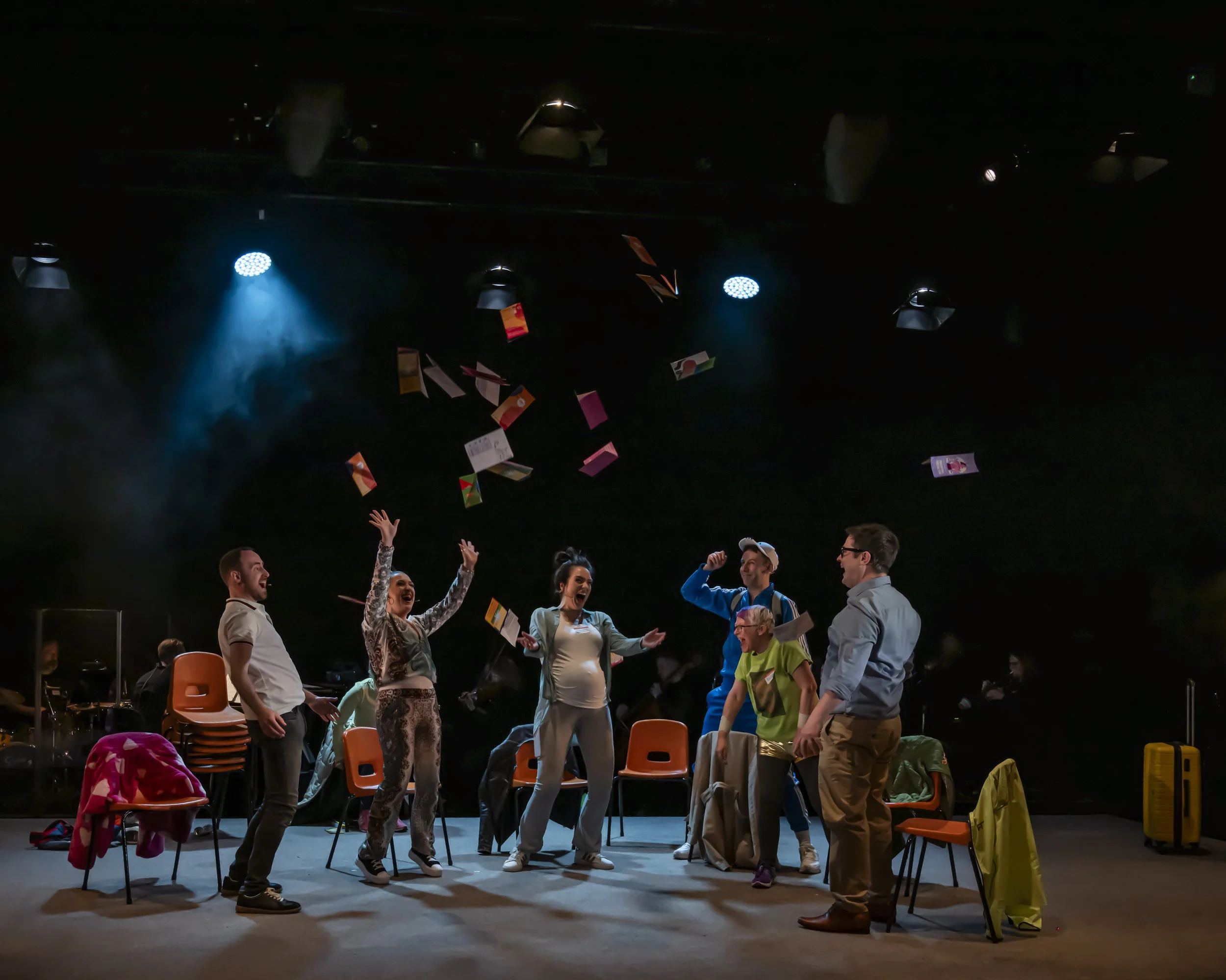 Group of people on a stage celebrating with raised hands, colorful confetti falling, chairs and costumes around, dark background with stage lights.