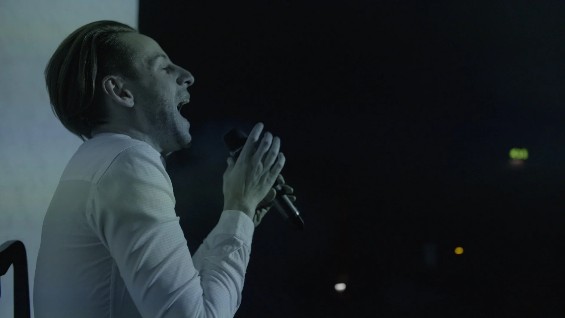 A man with light-colored hair singing into a microphone in a dark room with some lights in the distance.