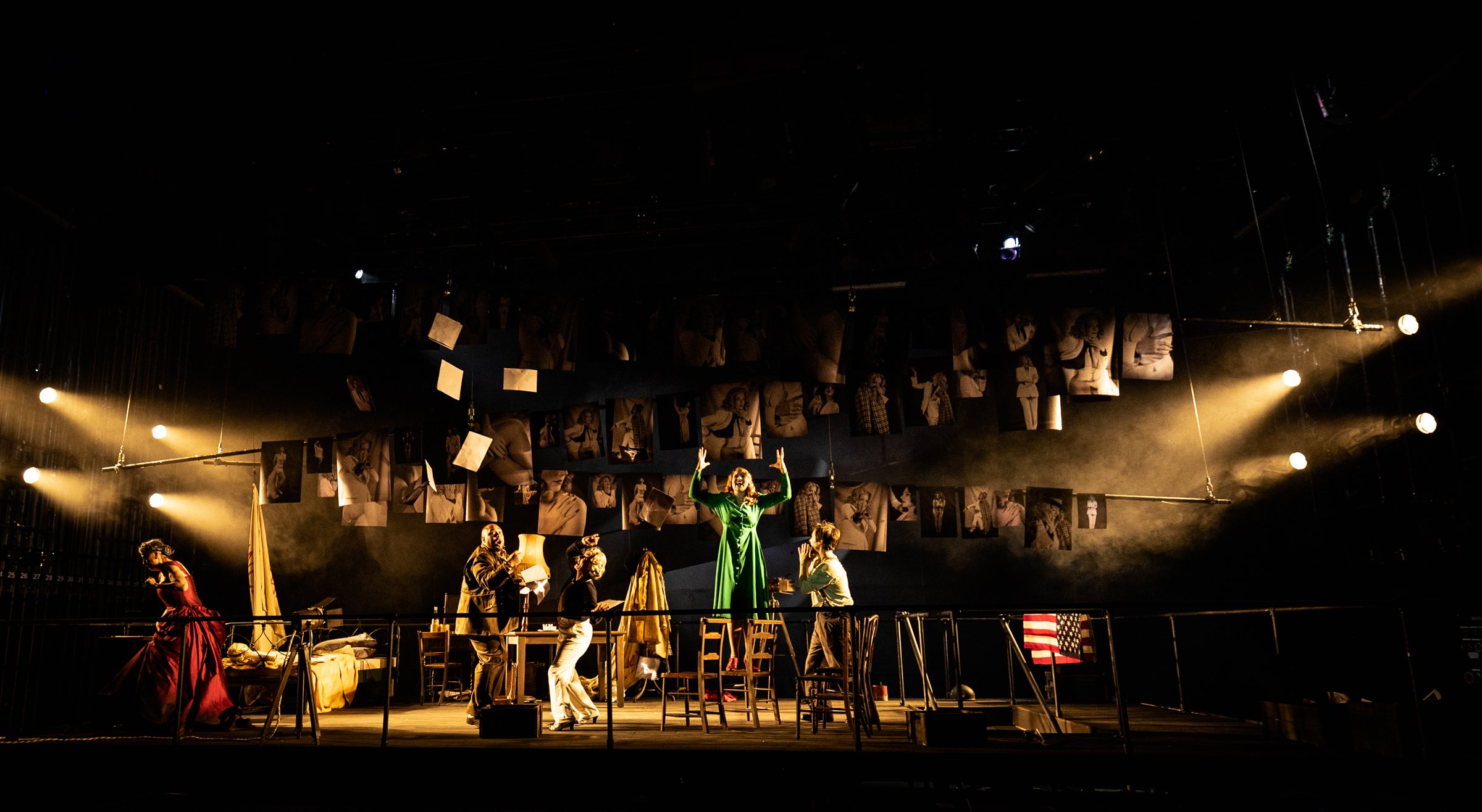 Theatrical stage scene with actors in period costumes, a woman in green center stage, surrounded by vintage furniture, photos hanging above, and dramatic lighting.