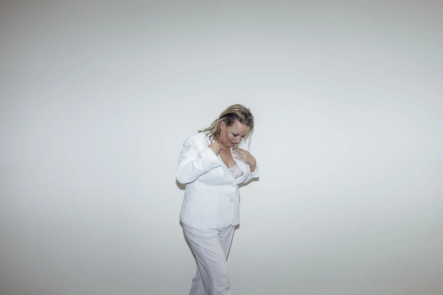 A woman with blonde hair in a white suit looking down, standing against a plain white background.