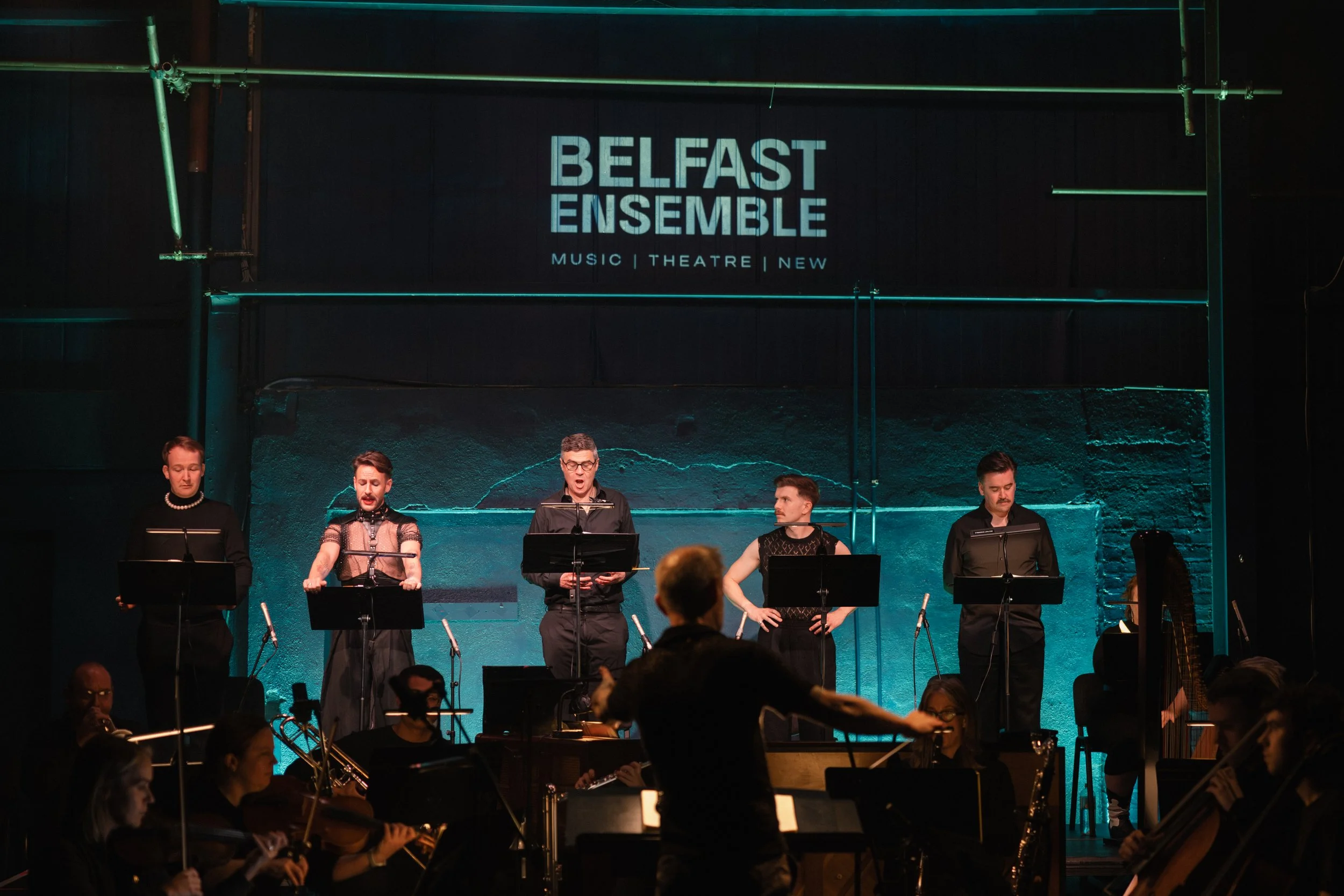 A group of musicians performing on stage during a concert at Belfast Ensmble, with some reading sheet music and others playing instruments, illuminated by stage lighting.
