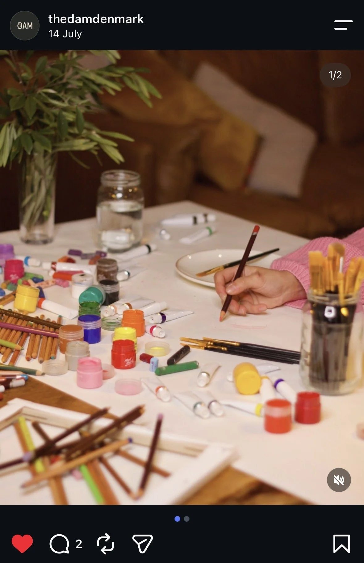 A person drawing on paper with colored pencils, surrounded by various art supplies, including paint pots, brushes, and markers, on a table, with a vase of greenery in the background.