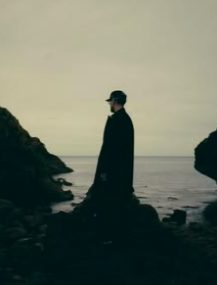 A person standing on rocks by the ocean, facing the water, wearing a hat and dark clothing, with cliffs on either side under an overcast sky.