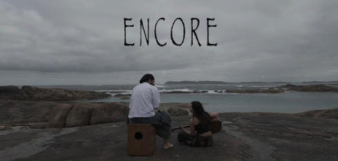 Two people sitting on a rocky shoreline in front of a body of water, under a cloudy sky, with the word 'ENCORE' floating above them.