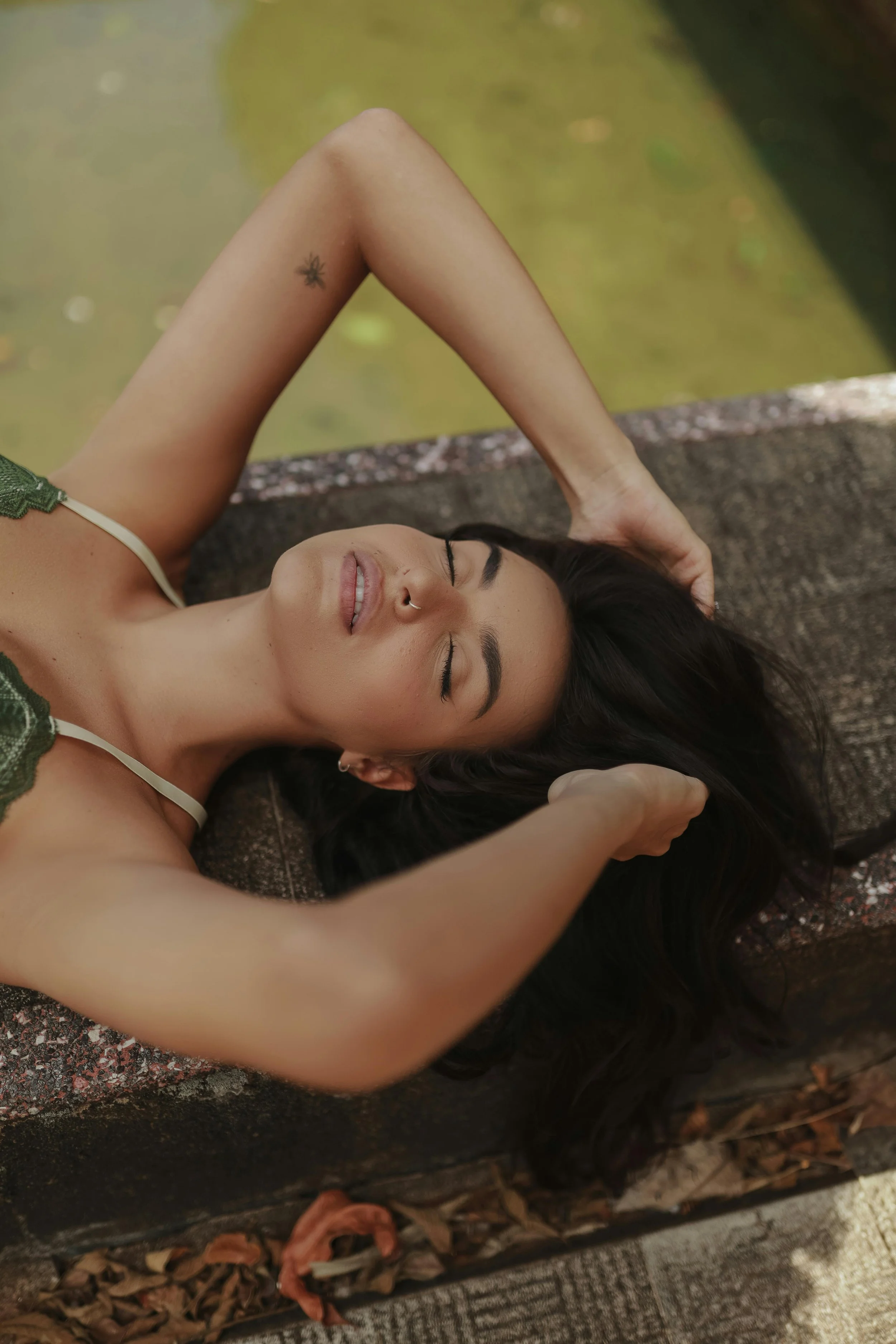 A woman with long dark hair and a nose ring is lying on her back with her eyes closed and a relaxed expression. She has her left arm raised above her head and her right arm extended to the side. She is dressed in a green top with lace details and is outdoors next to a water feature, surrounded by fallen leaves.
